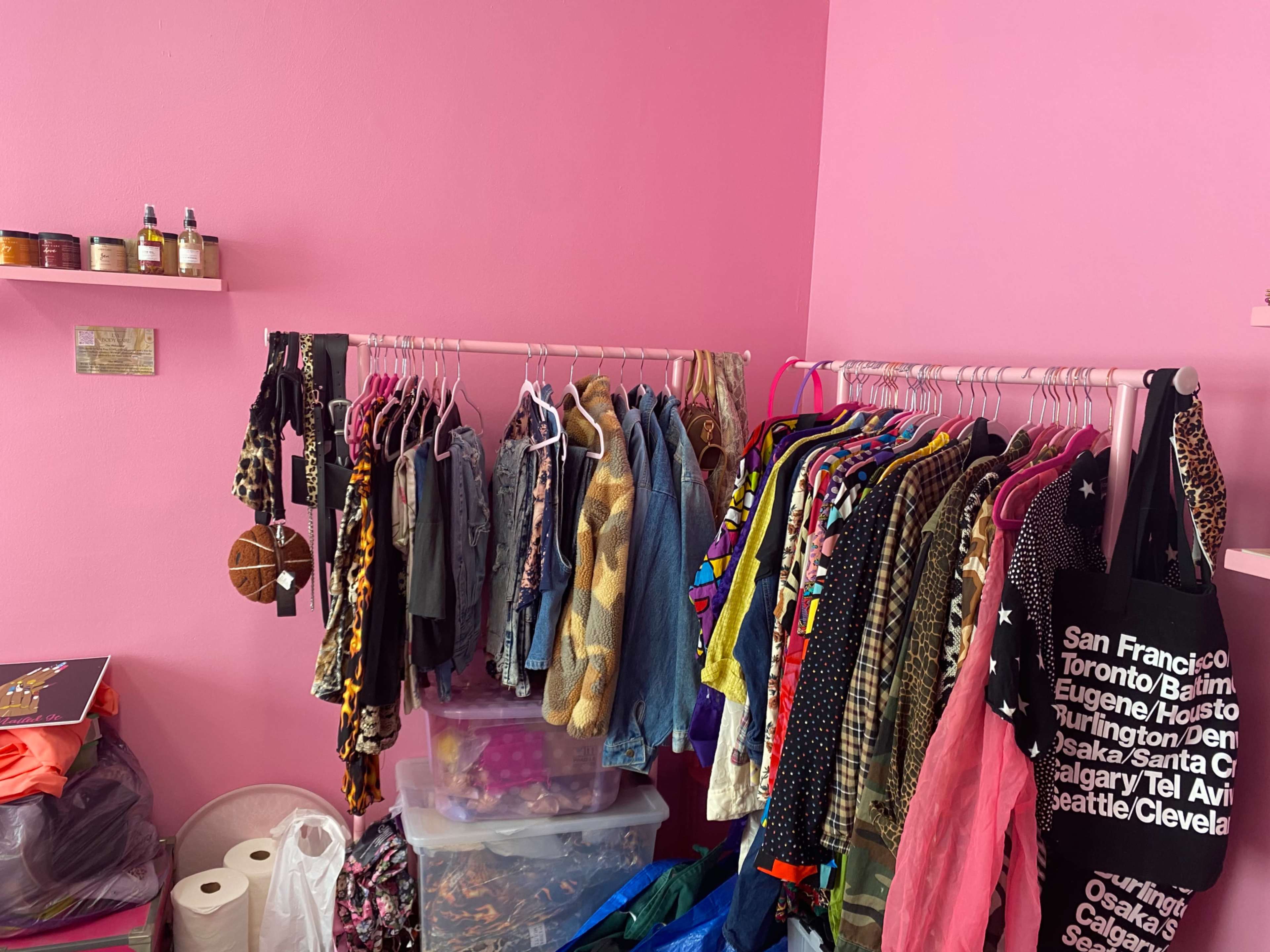 The image shows a room with pink walls featuring two clothing racks filled with various garments, alongside bins and bags of additional clothing items.