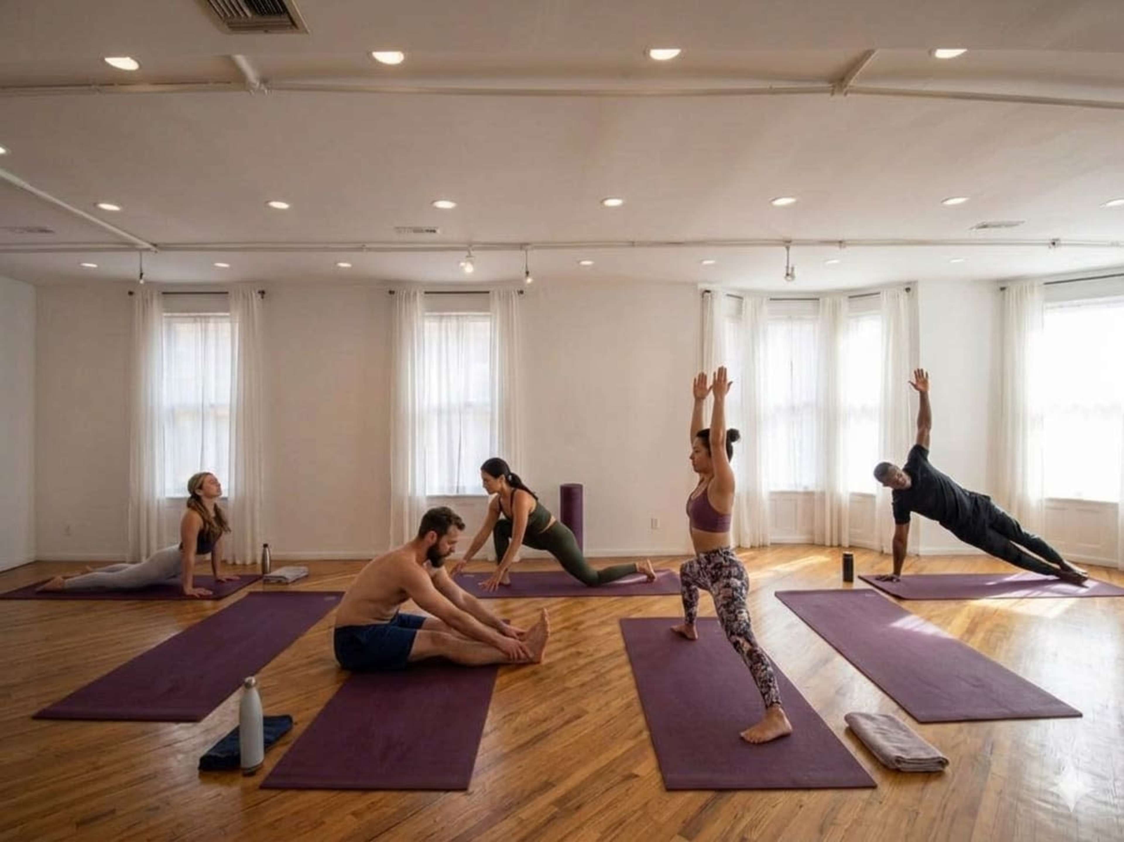 A group of five individuals practices yoga in a spacious studio with wooden floors and natural light coming through large windows.