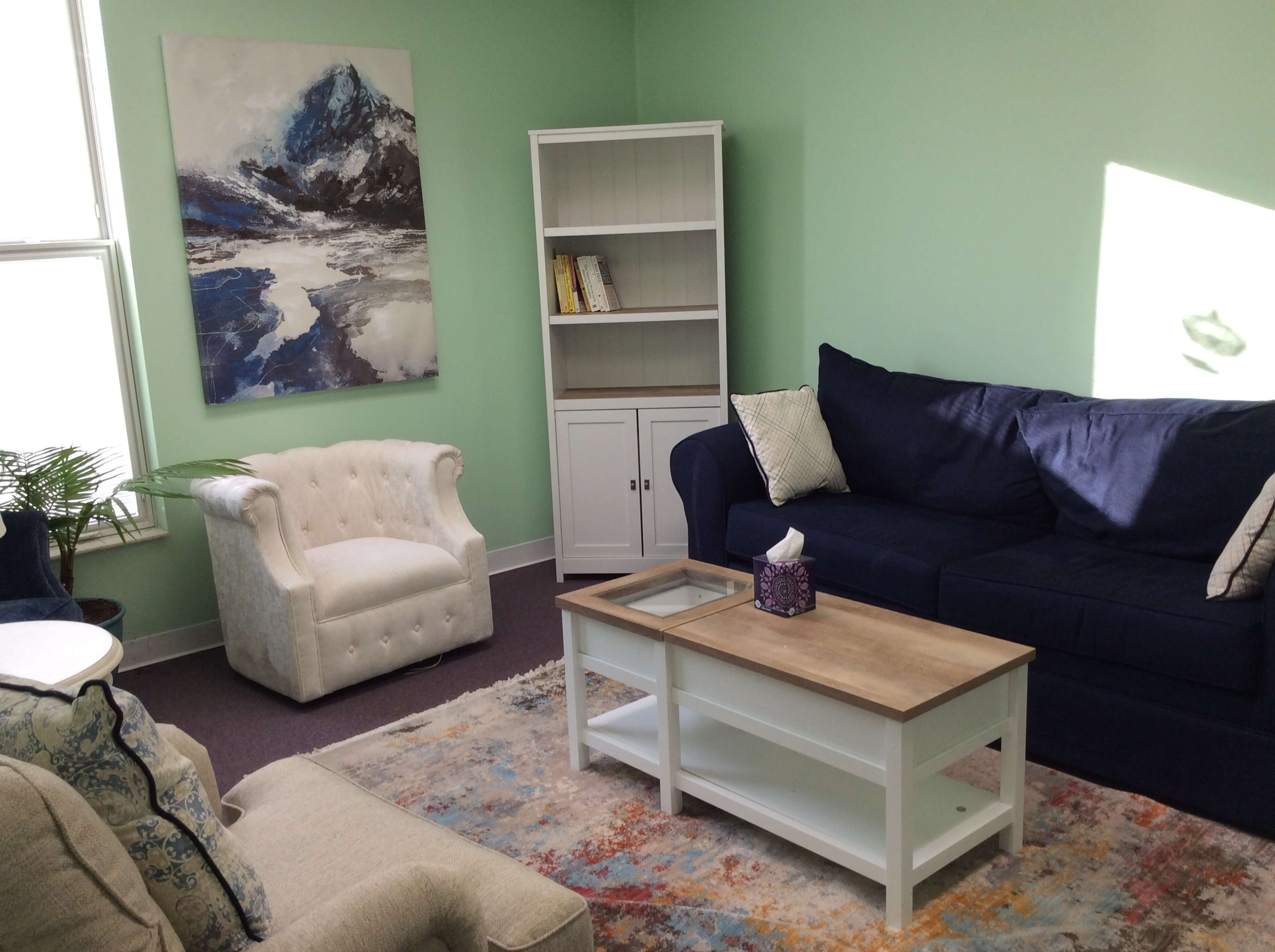 A brightly lit room with green walls, featuring a blue couch, a white tufted armchair, a wooden coffee table, a bookshelf, and a mountain landscape painting on the wall.