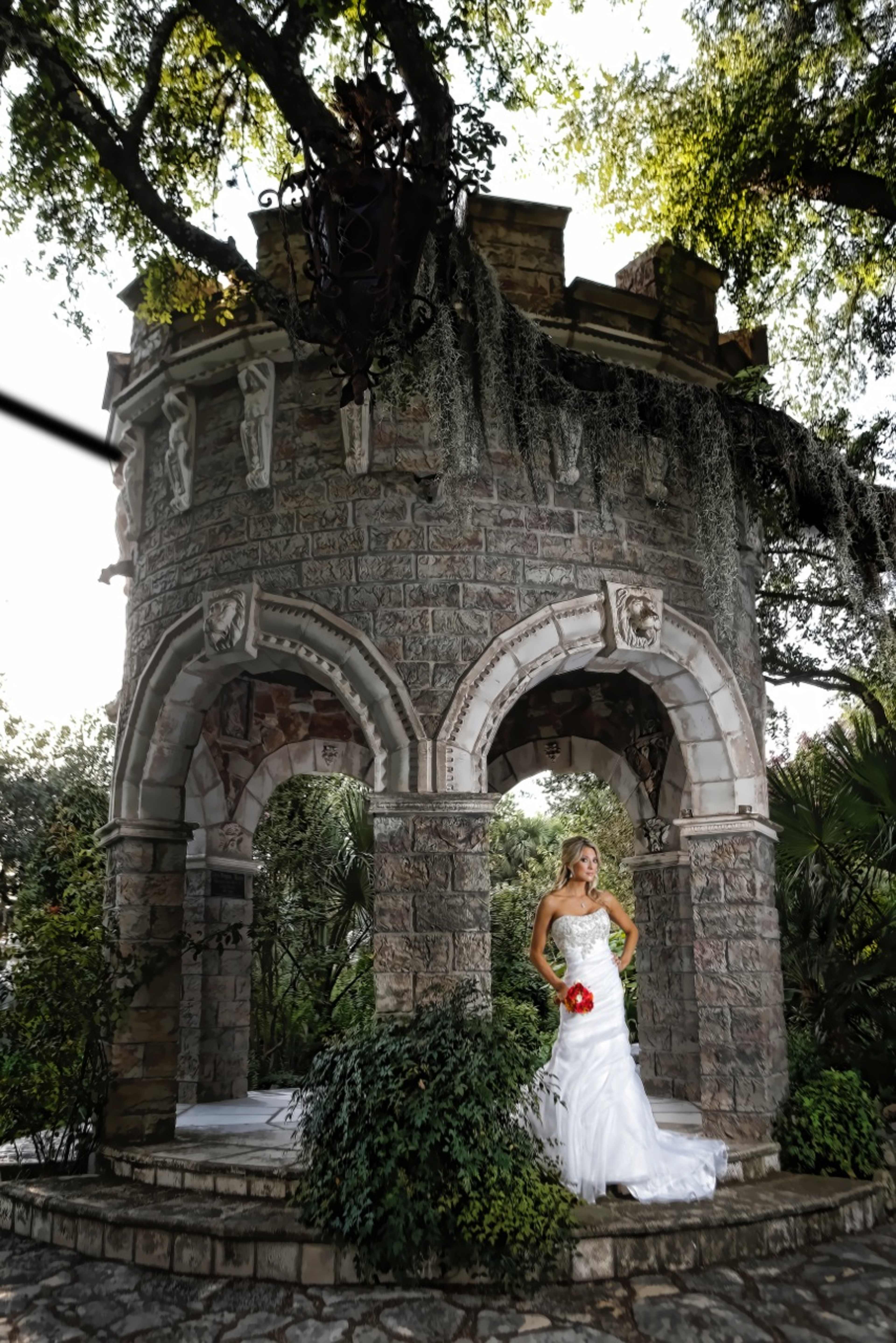 A bride in a white dress holds a red bouquet while standing in front of a stone gazebo surrounded by lush greenery.