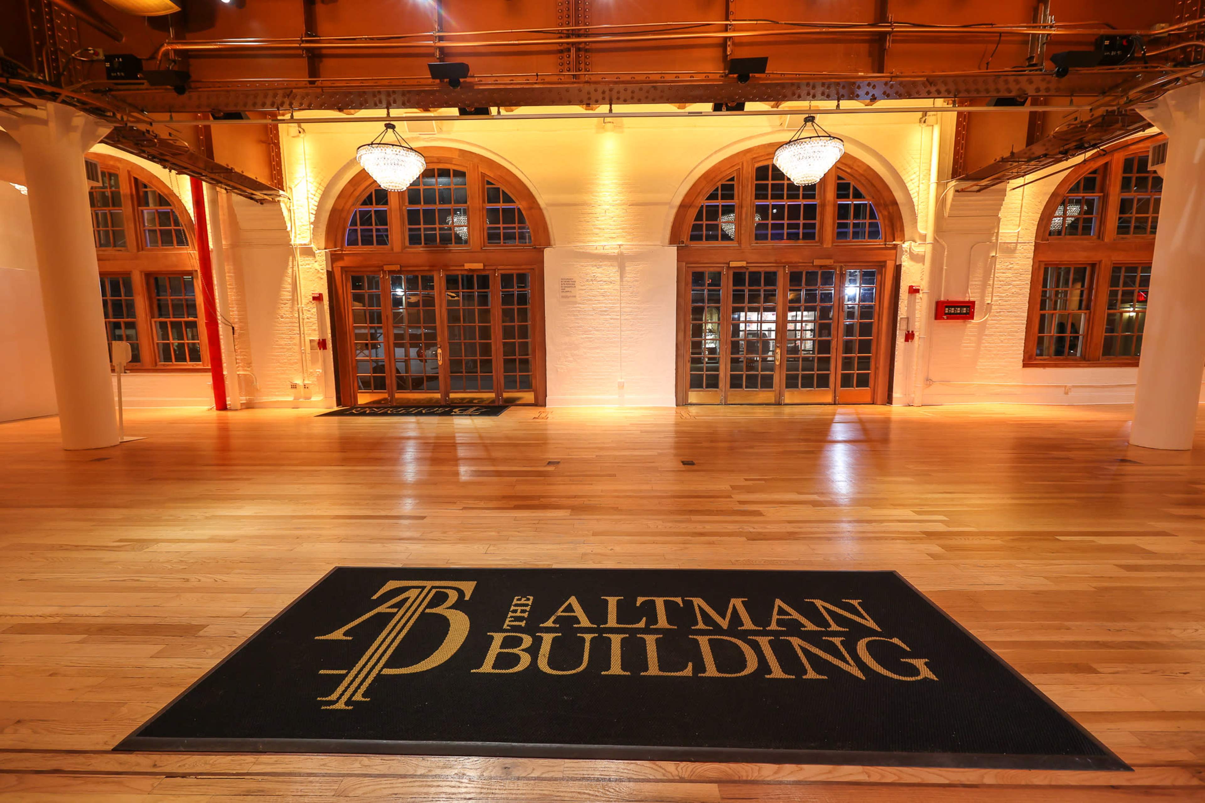 The interior of the Altman Building features wooden flooring, large arched windows, and a prominent entrance mat displaying its name.