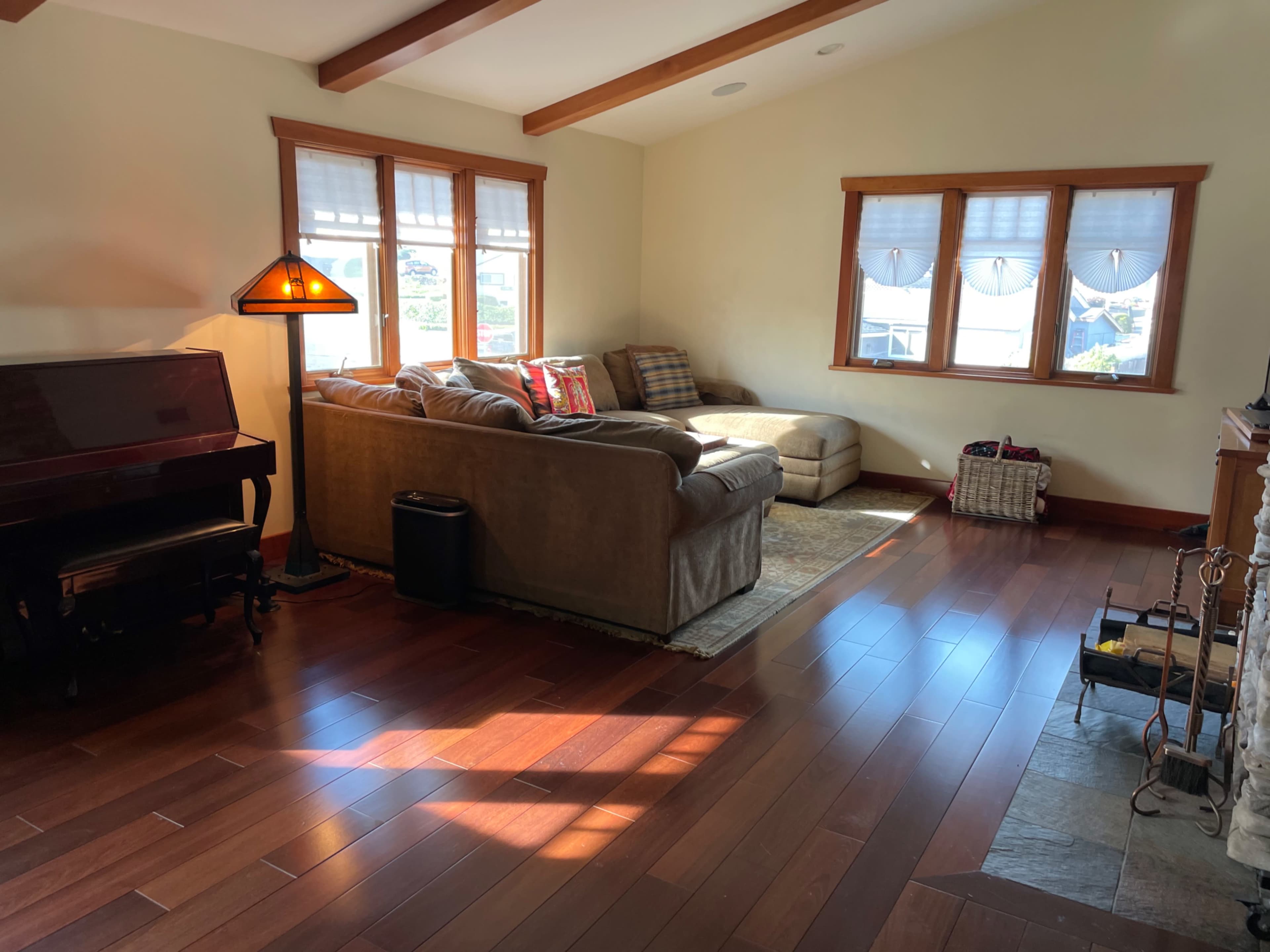 The living room features a sofa, a chaise lounge, a piano, and large windows that illuminate the hardwood floor.