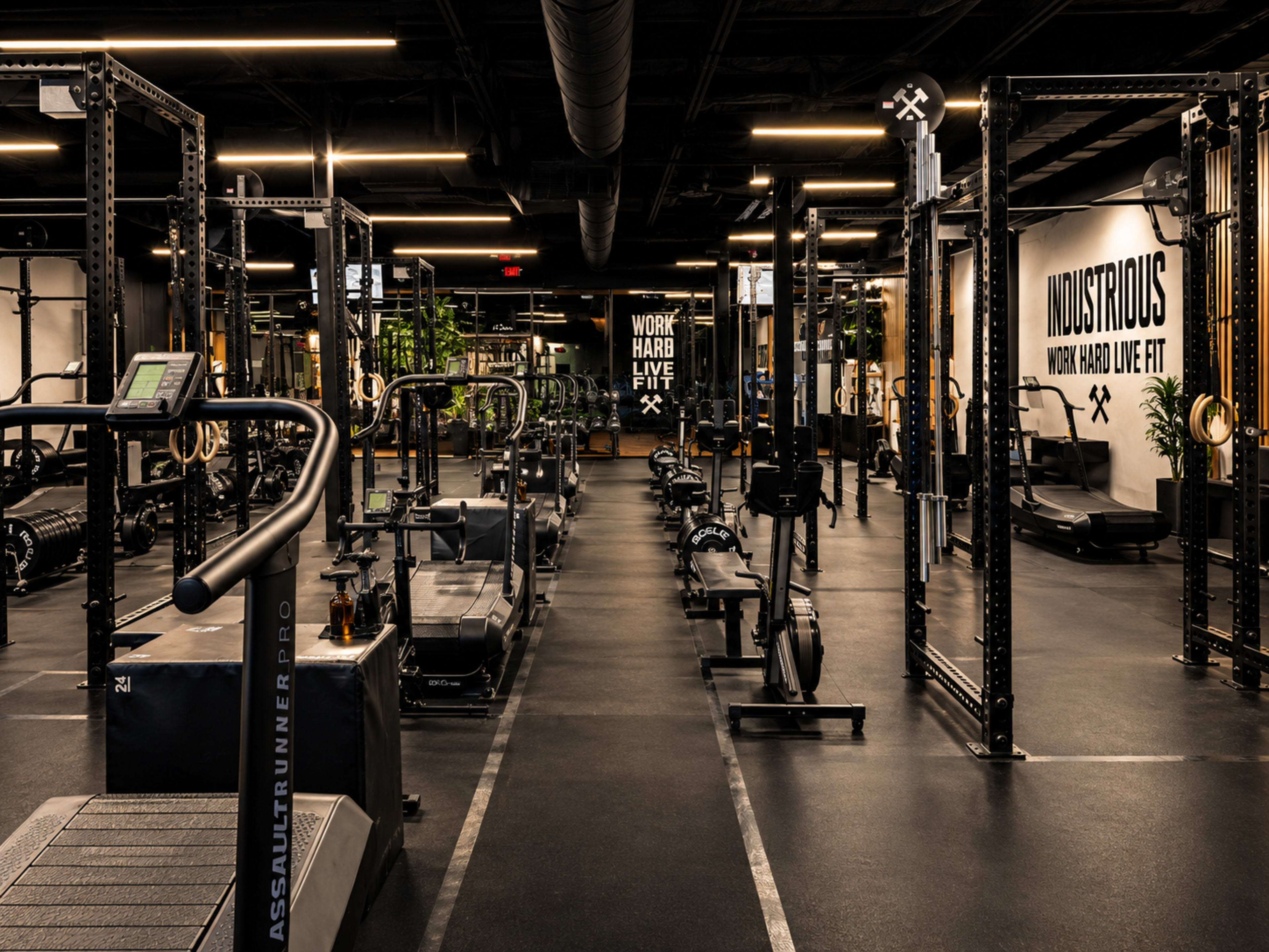 The image shows a well-equipped gym with various workout machines, including treadmills, rowing machines, and weightlifting stations, all arranged in an organized manner.