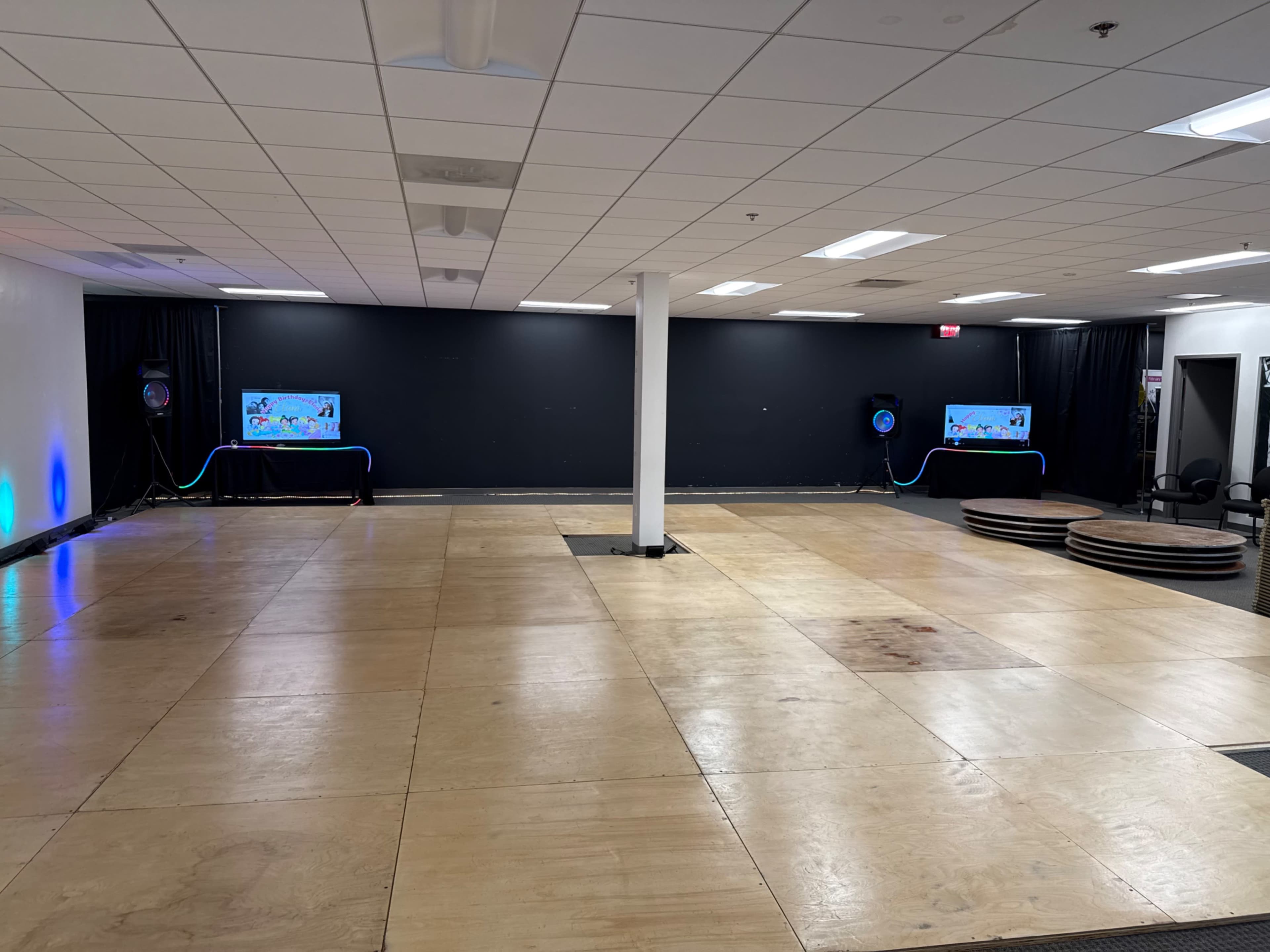 The image shows a spacious indoor area with wooden flooring, two screens displaying visuals, and a central pillar, all under fluorescent lighting.