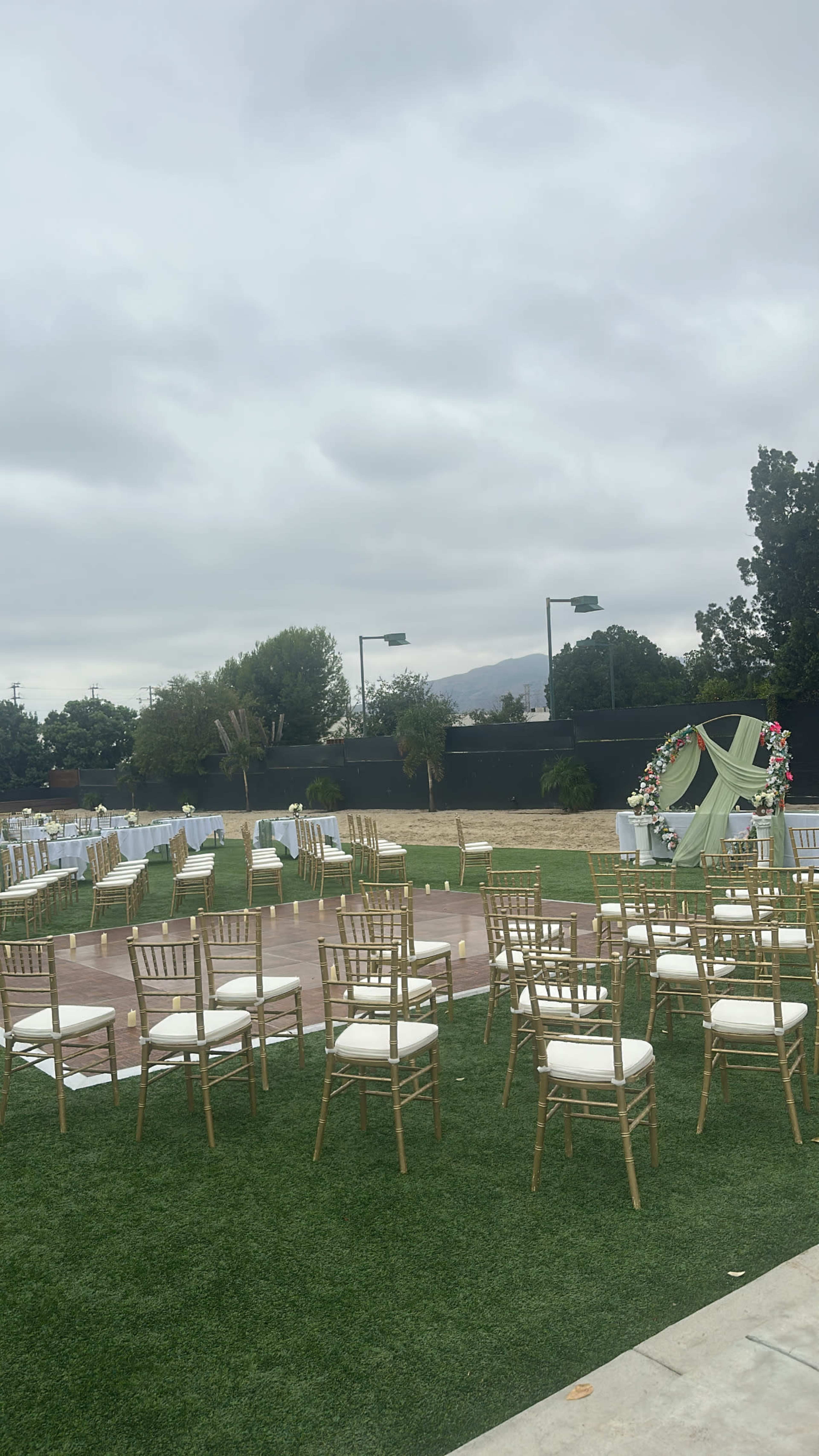 The image shows a wedding setup with rows of golden chairs arranged on a lawn beneath a cloudy sky, featuring a floral arch at one end.