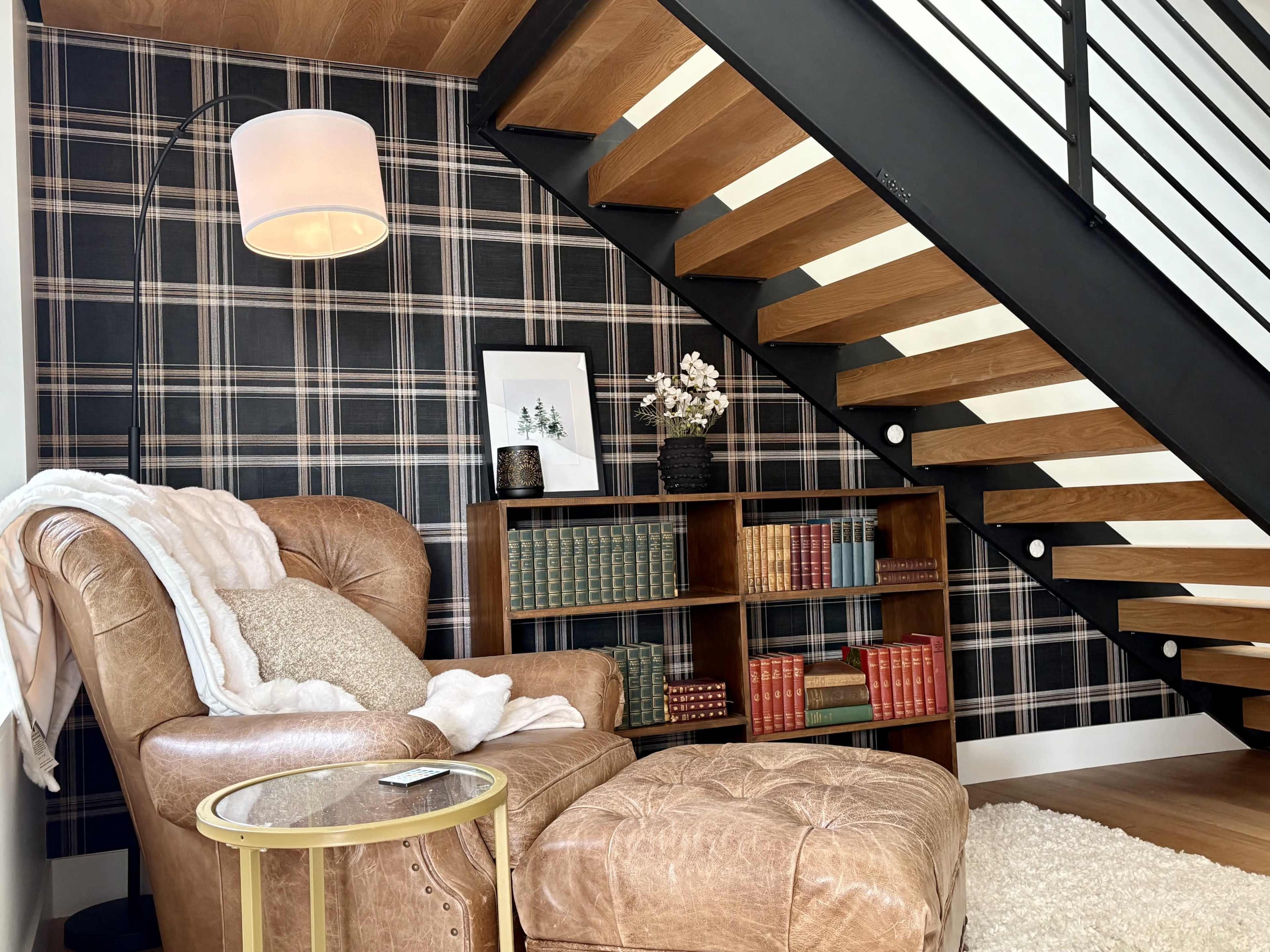 The image shows a cozy reading nook featuring a leather armchair with a throw blanket, a small round table, and a bookshelf filled with books, all situated under a staircase with patterned wallpaper.