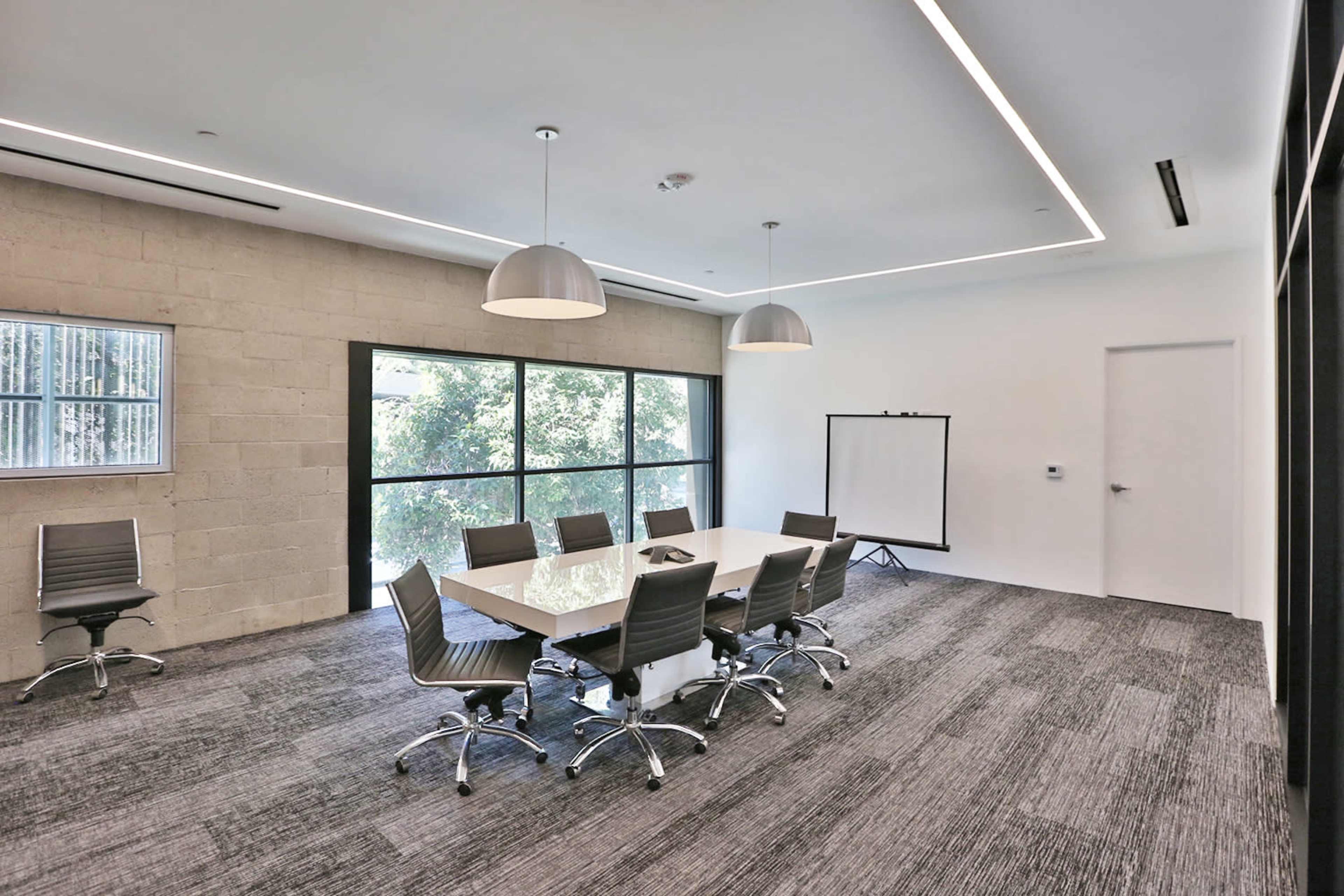 A modern conference room with a rectangular table surrounded by eight rolling chairs, large windows letting in natural light, and a whiteboard on a stand.