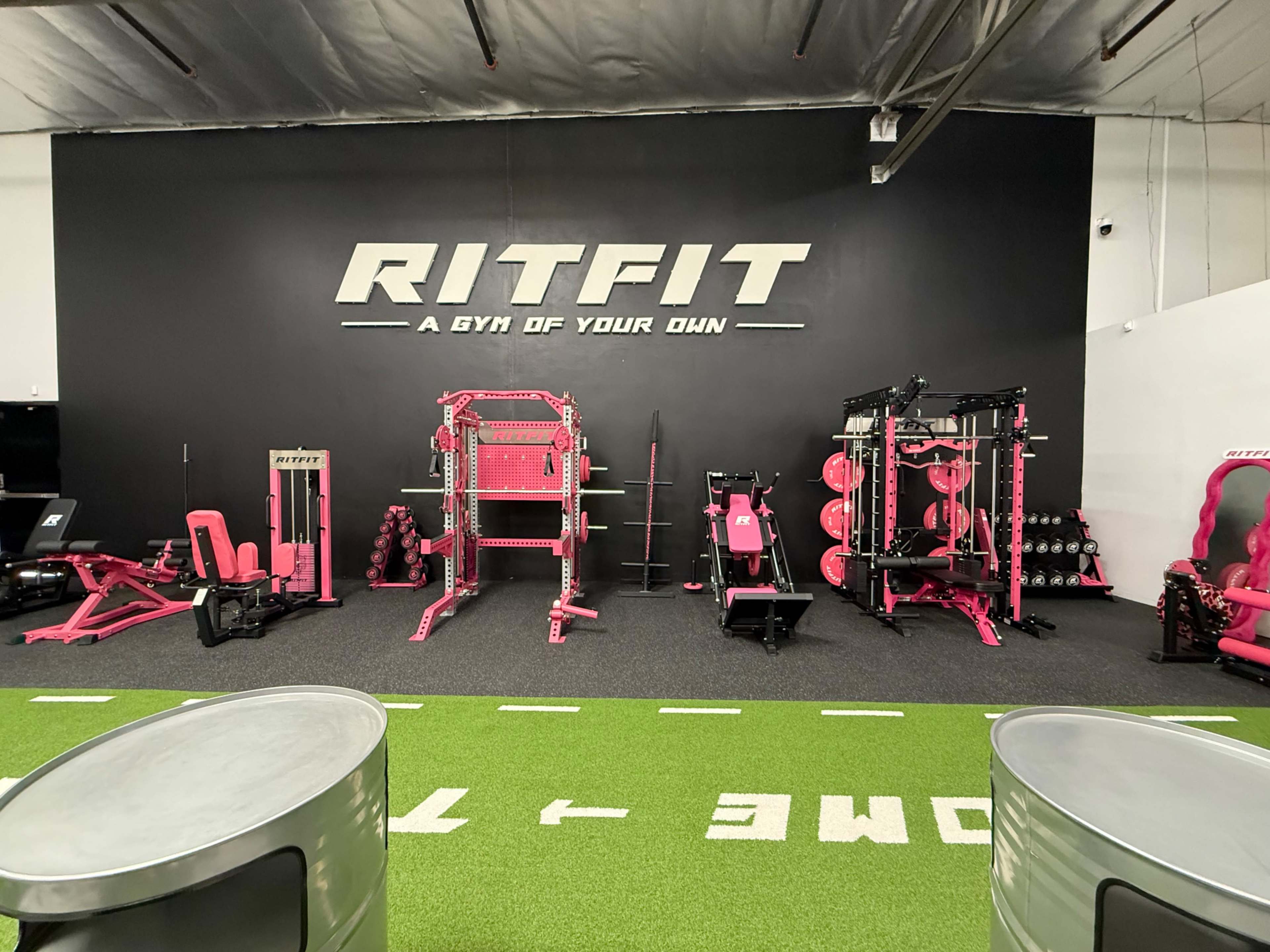 The image shows a modern gym interior with pink workout equipment and weights set against a black wall that features the name "RITFIT."