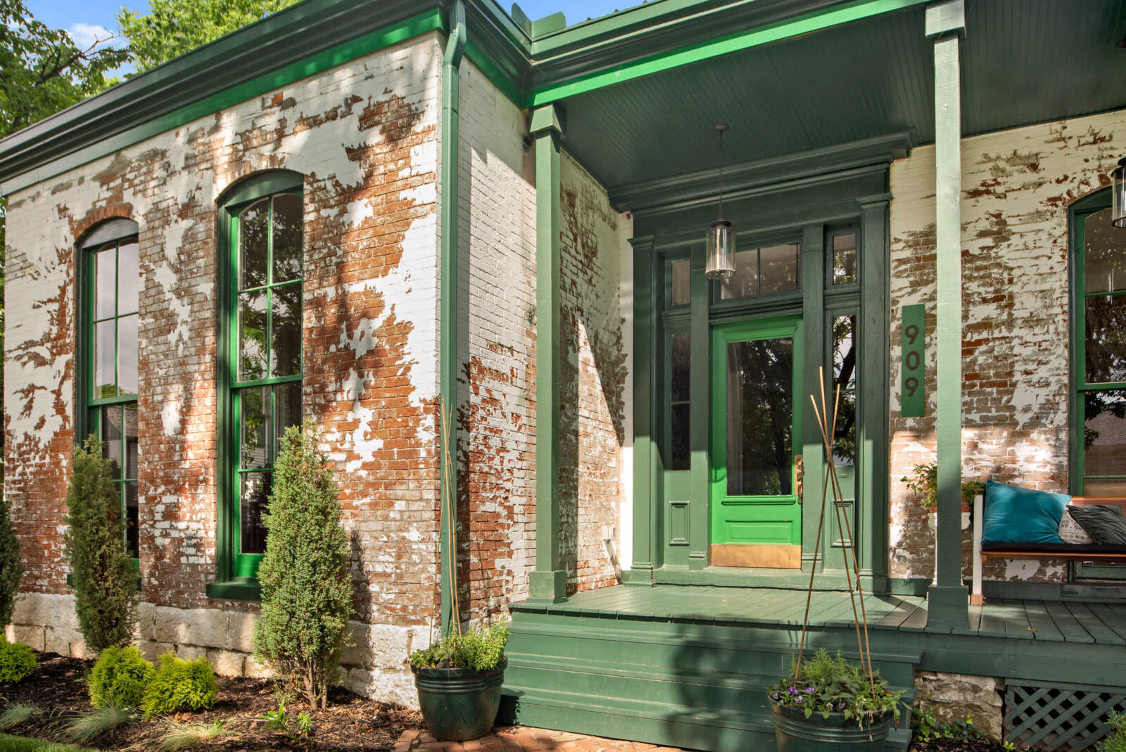 A house with a brick exterior featuring peeling paint, green trim, and a front porch with potted plants.