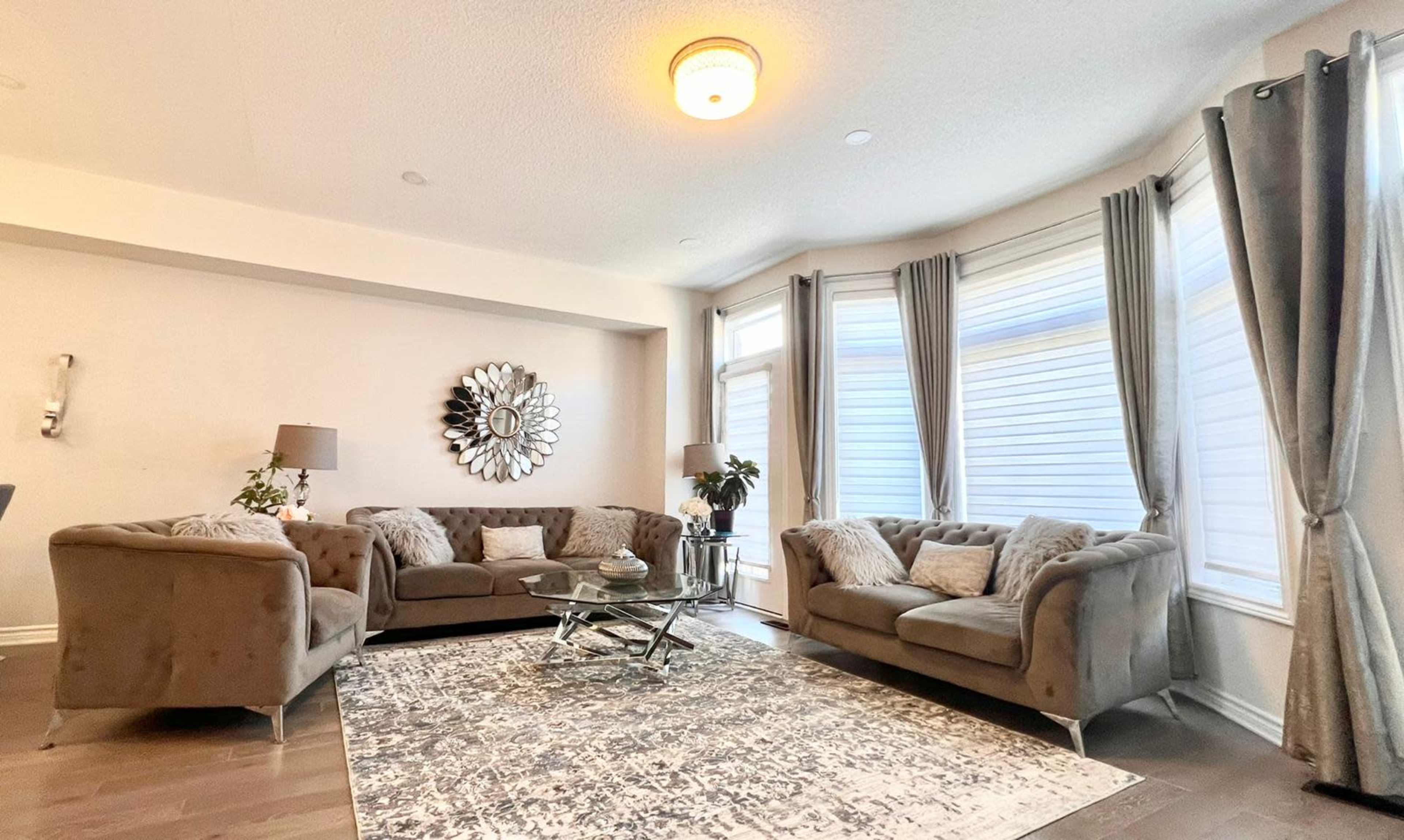 A modern living room features a sectional sofa, a glass coffee table, decorative pillows, and large windows with sheer curtains.