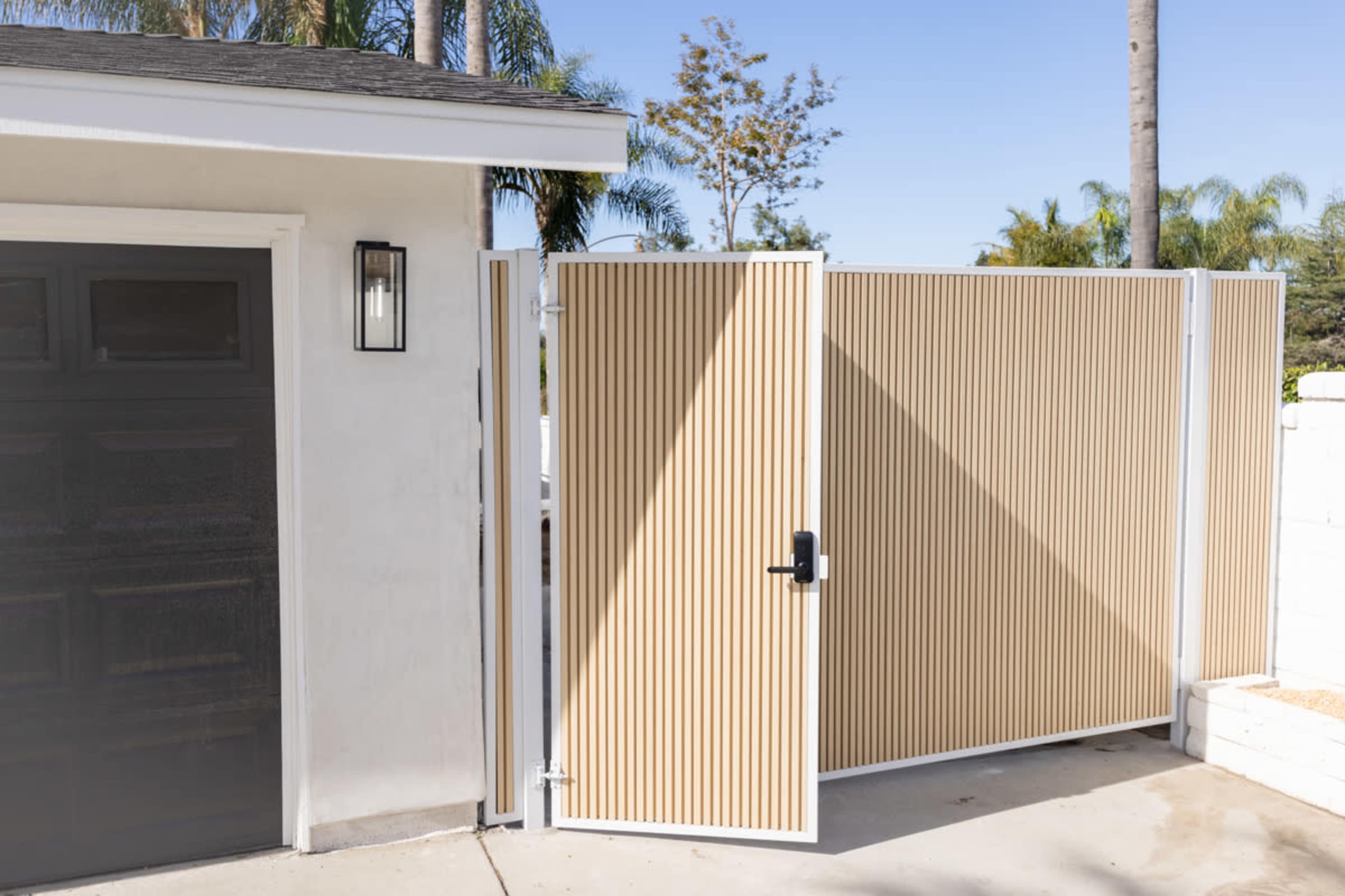 A beige horizontal slatted gate stands open next to a dark garage door, with palm trees visible in the background.