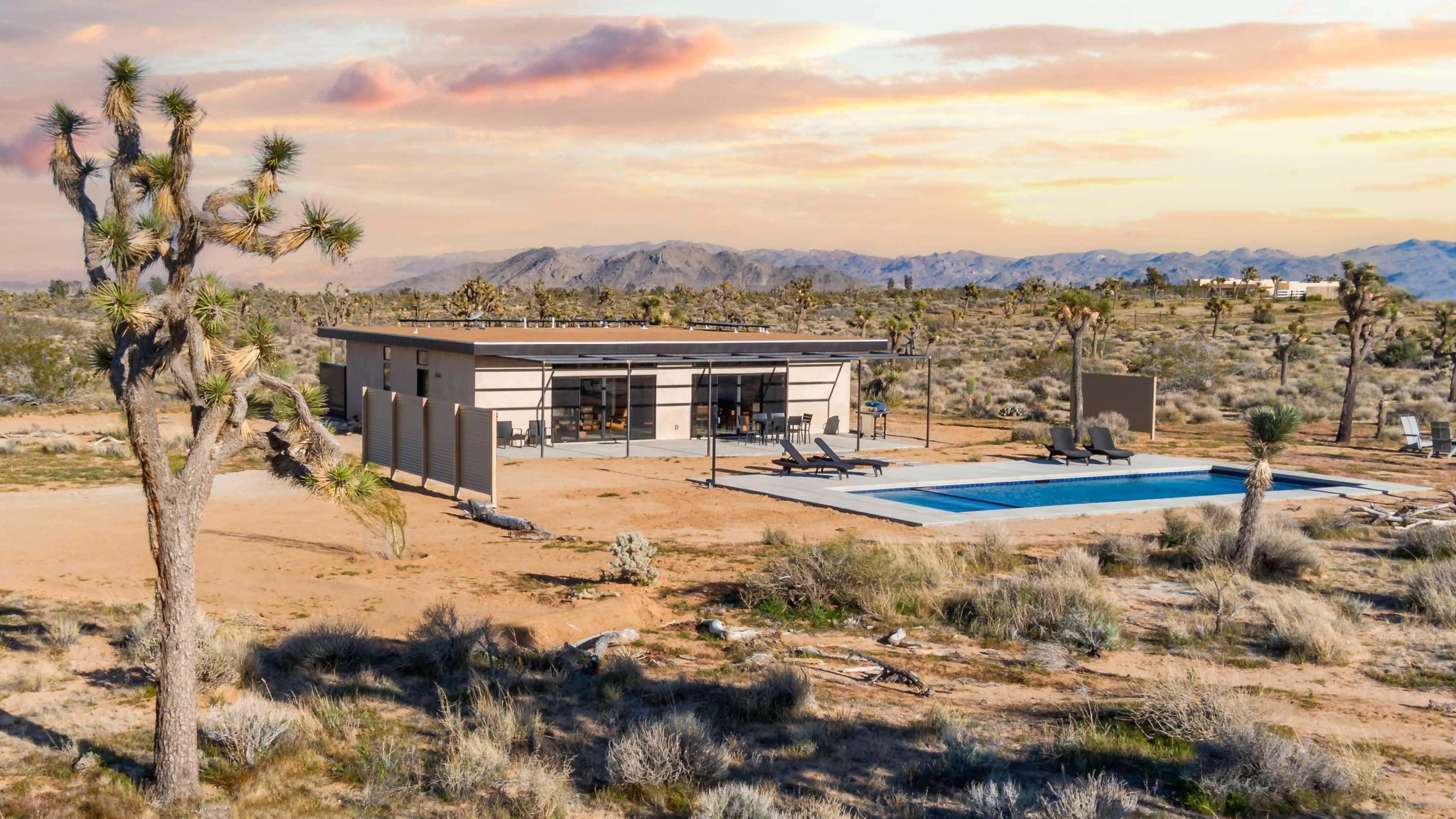 A modern house with large windows and a pool is set in a desert landscape surrounded by sparse vegetation and distant mountains.