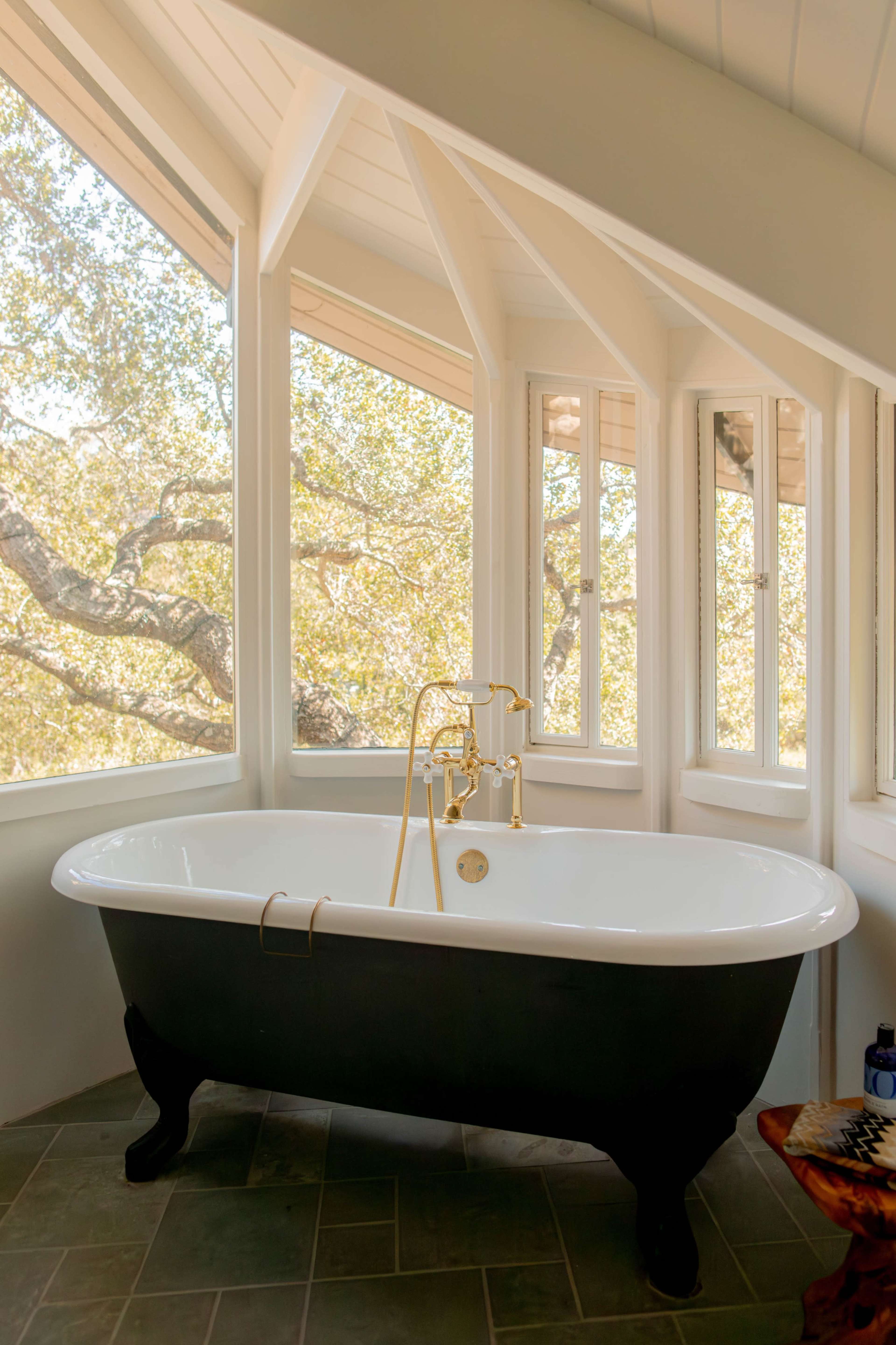 A black freestanding bathtub with gold fixtures is positioned near large windows that offer a view of outdoor trees in a well-lit bathroom.