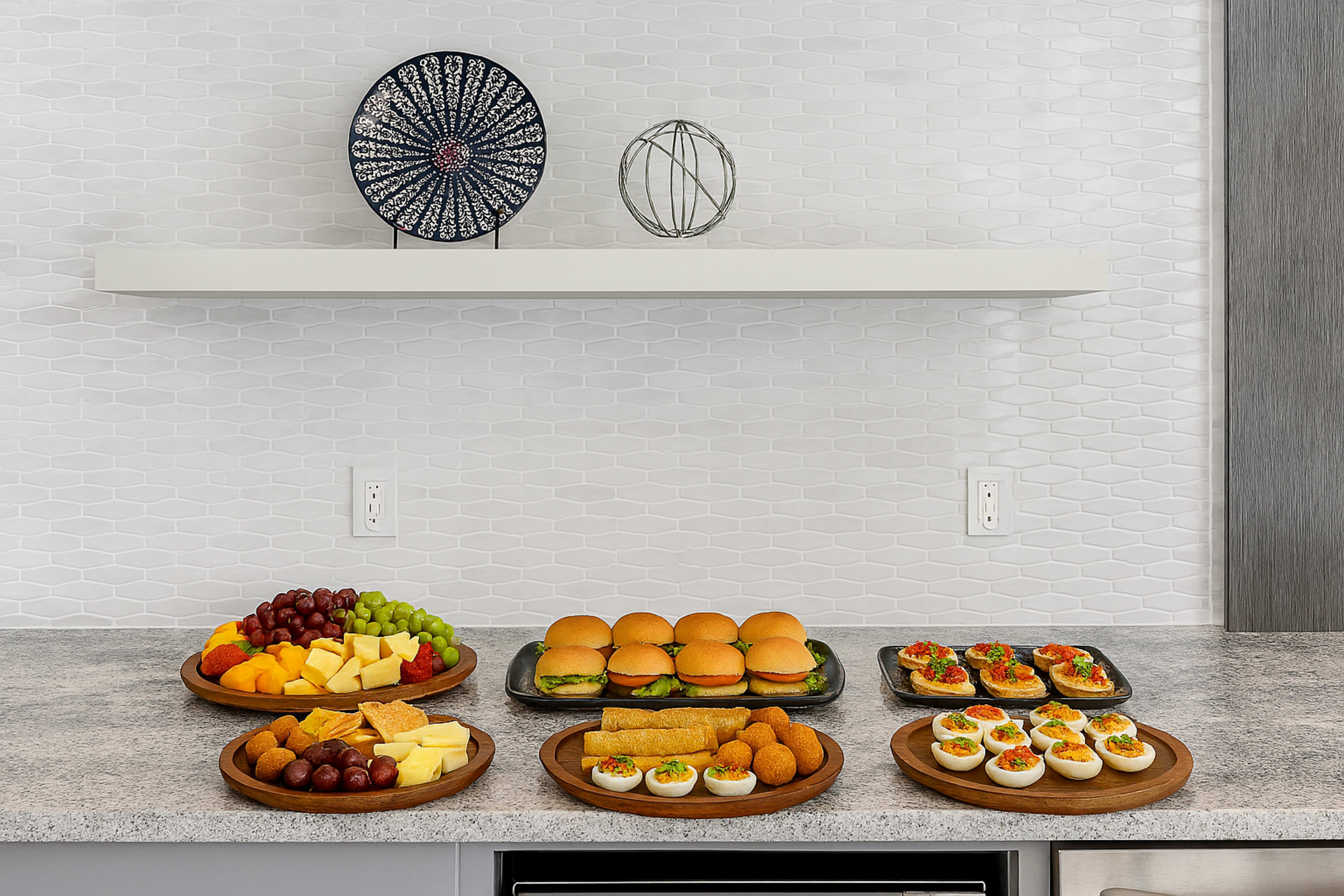 The image features a buffet arrangement on a countertop with various food platters, including fruits, cheese, rolls, and appetizers.