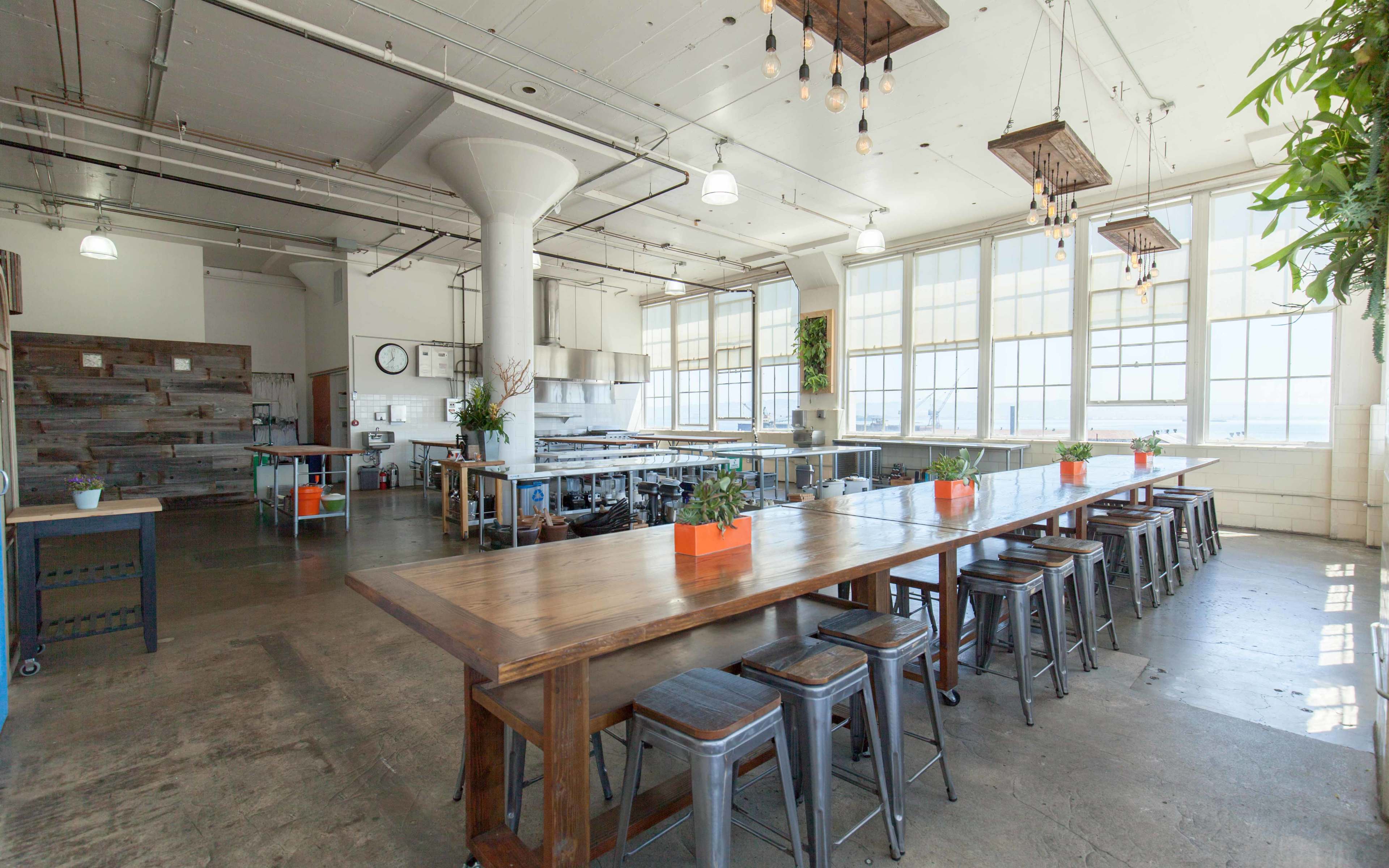 A spacious, modern kitchen with large windows, high ceilings, and a long wooden dining table surrounded by metal stools.