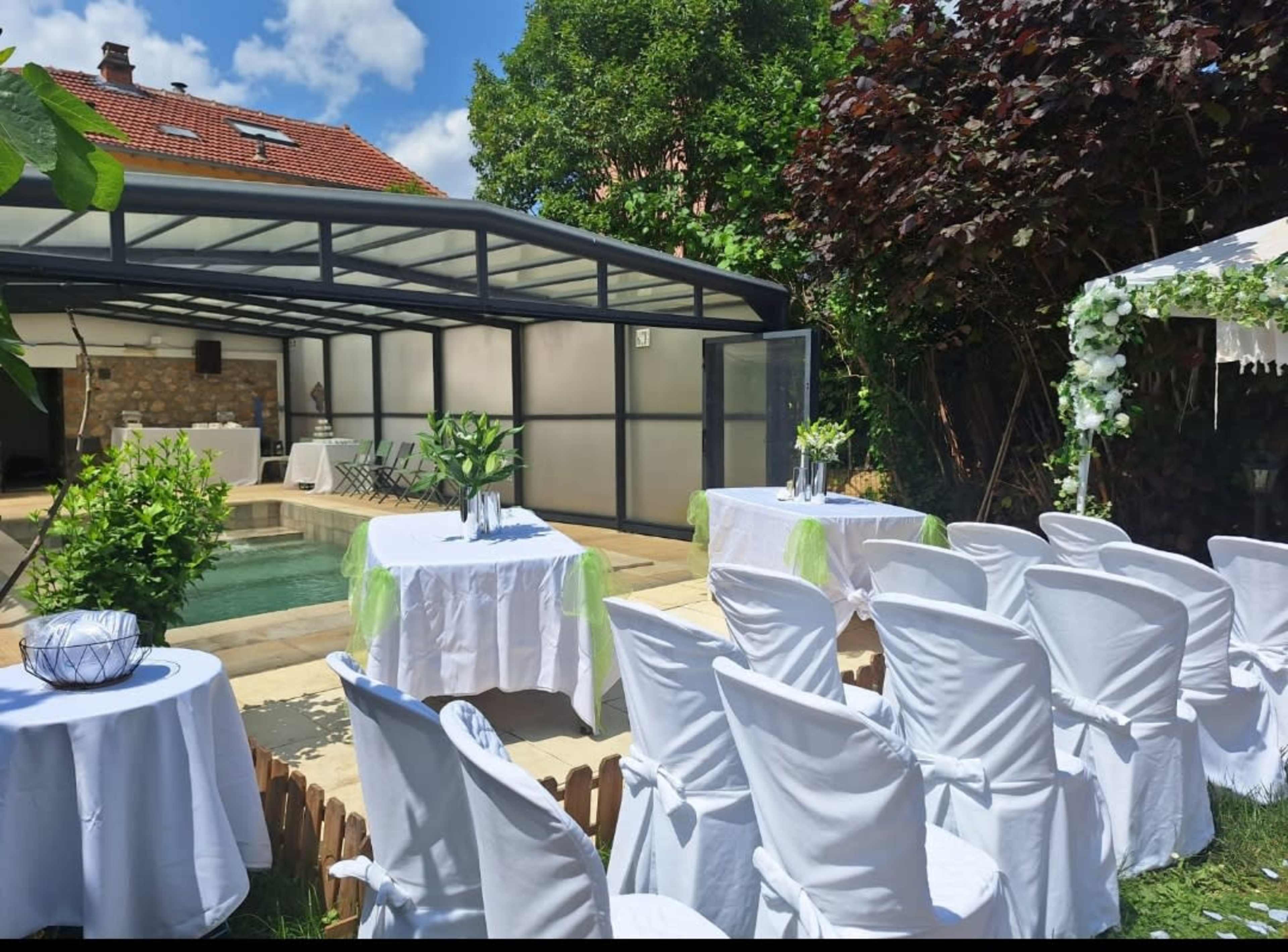 A backyard setting features a pool surrounded by white-clothed tables and chairs, with a gazebo adorned with decorations and greenery.