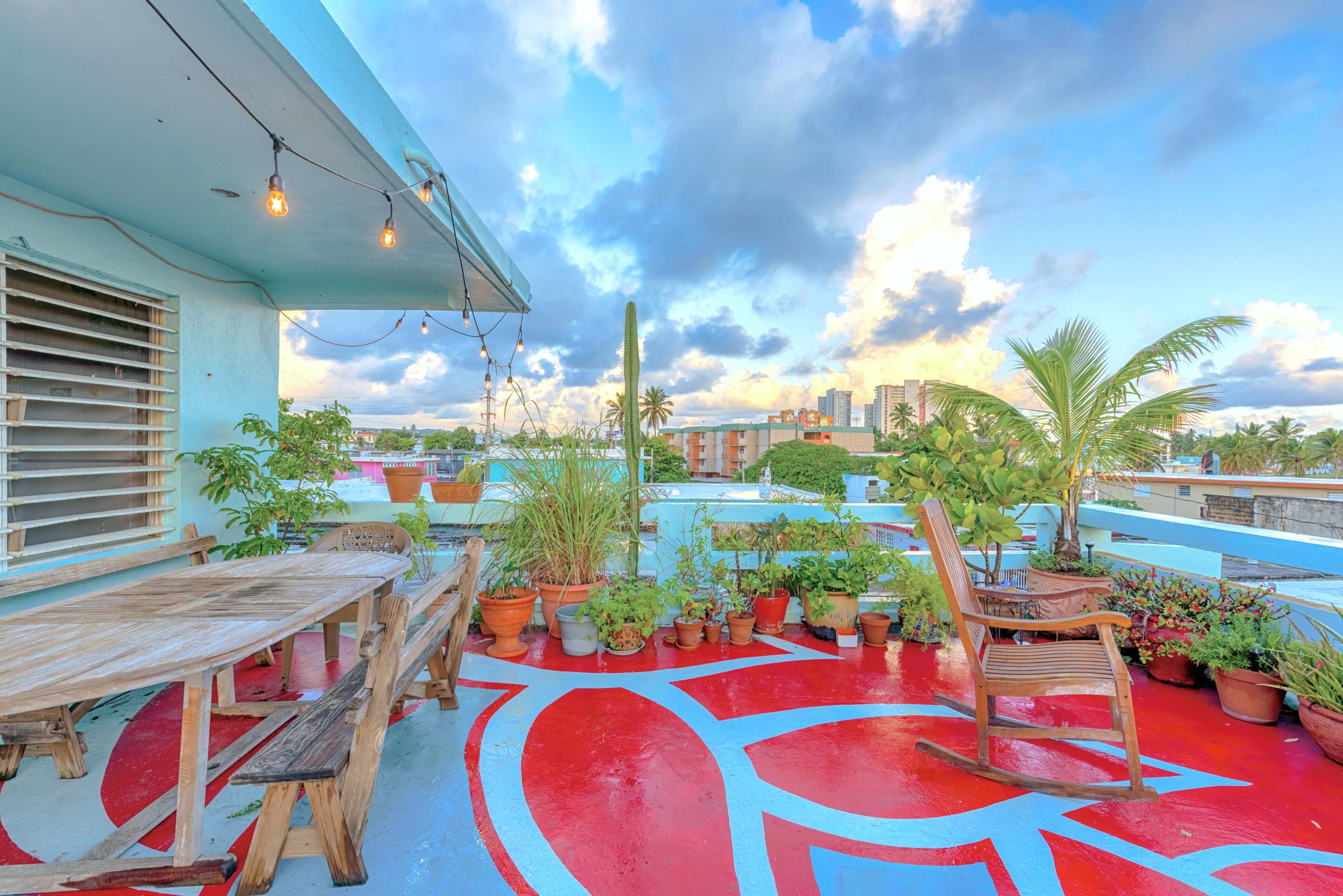 The rooftop terrace features a wooden table, chairs, and numerous plants in pots, with a colorful painted floor and a view of the city skyline.
