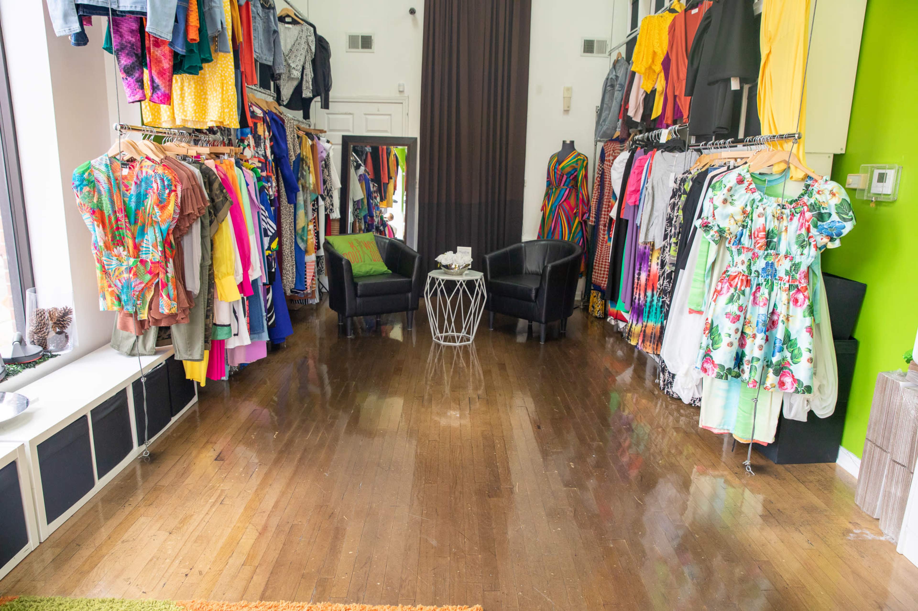 The image shows a clothing boutique with racks of colorful garments on either side and two black chairs facing a small white table in the center.