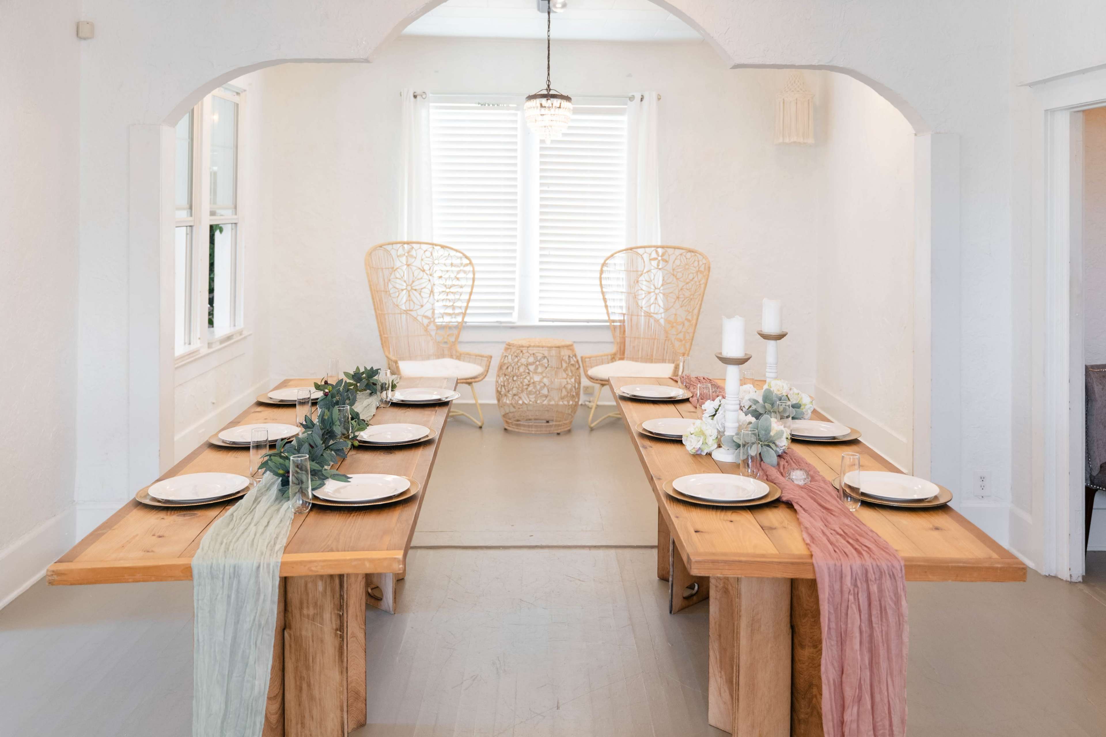 A minimalist dining room features two long wooden tables set with plates, surrounded by light-colored walls and framed by decorative chairs and a small table near a window.