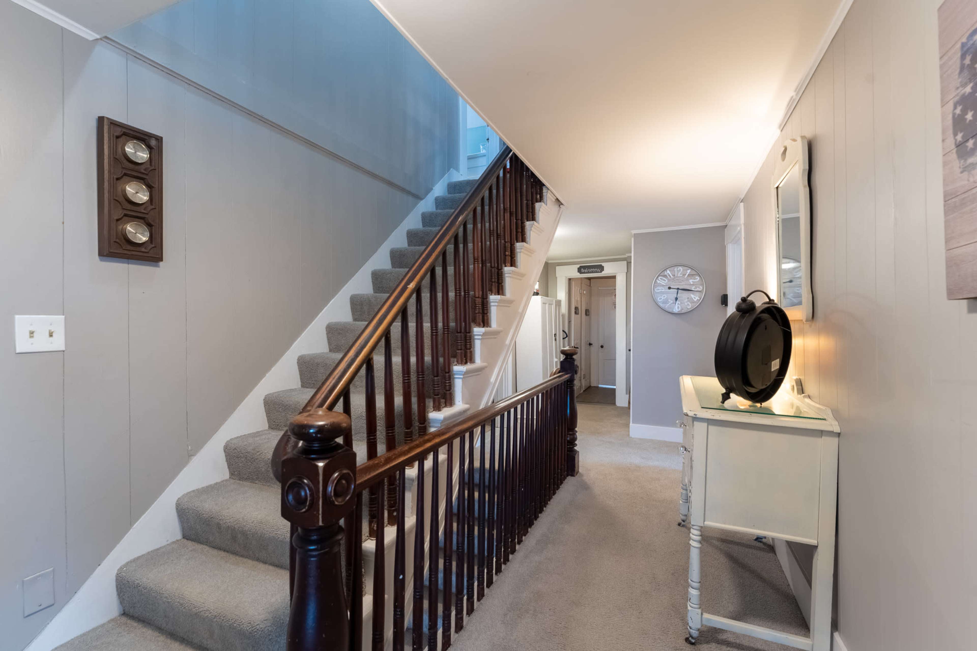 A staircase with wooden banisters leads to an upper level, flanked by gray walls and a small table with a round mirror on the landing.