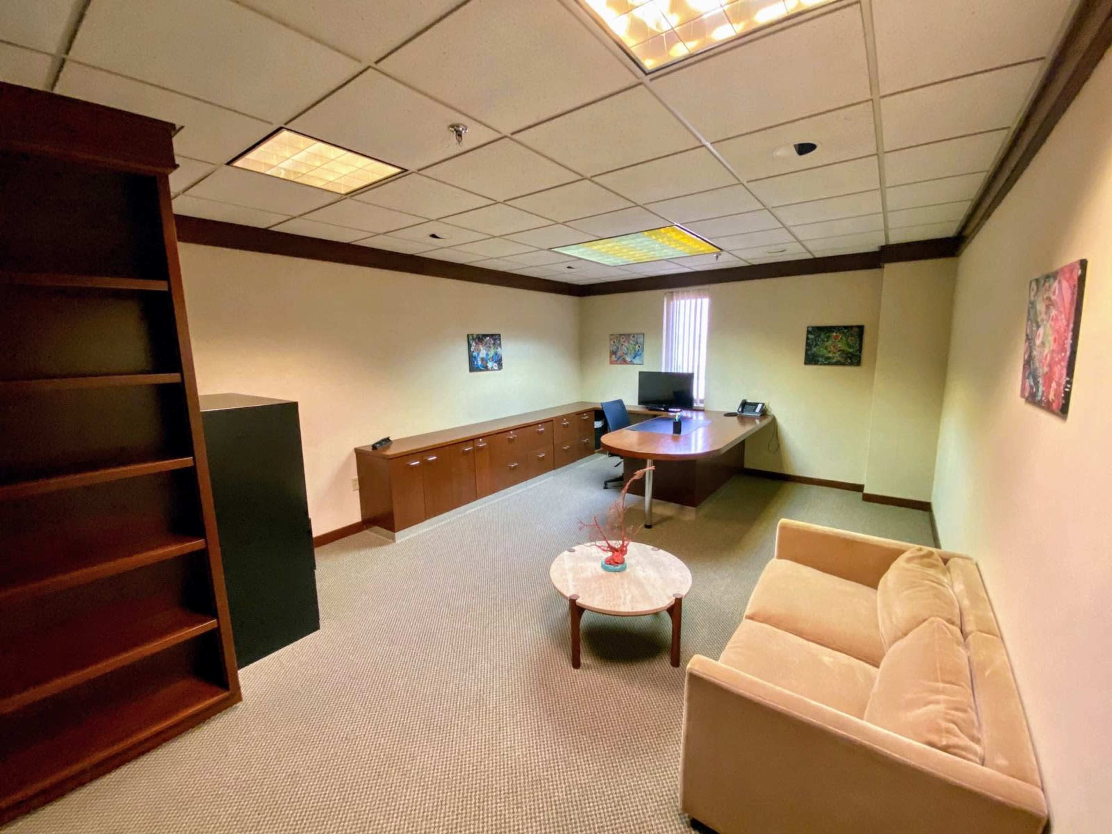 A sparsely furnished office with a desk, computer, bookshelf, and a sofa, featuring light-colored walls and a patterned carpet.