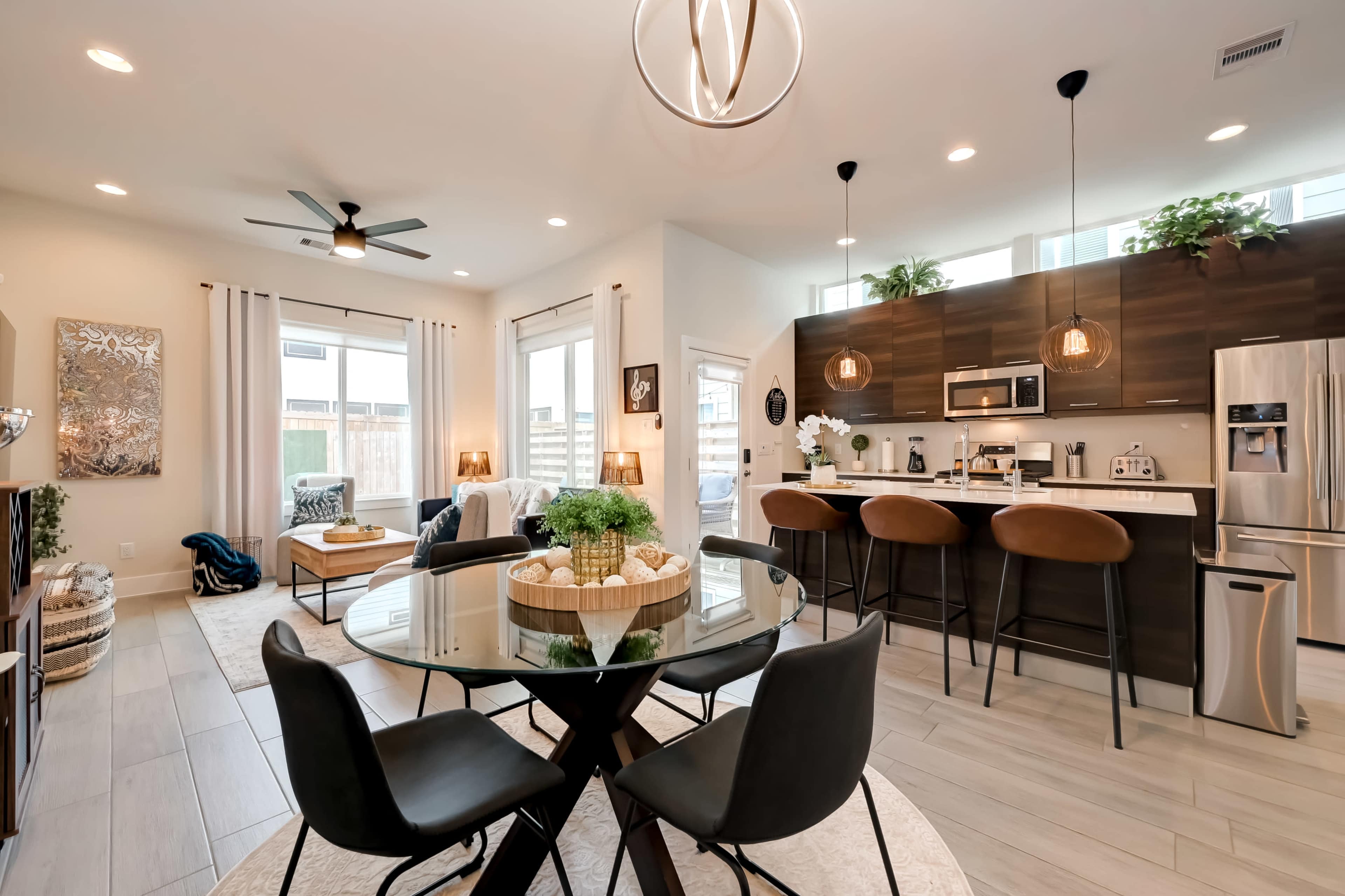 A modern kitchen and dining area with a circular glass table, four black chairs, and a kitchen island equipped with stainless steel appliances and pendant lighting.