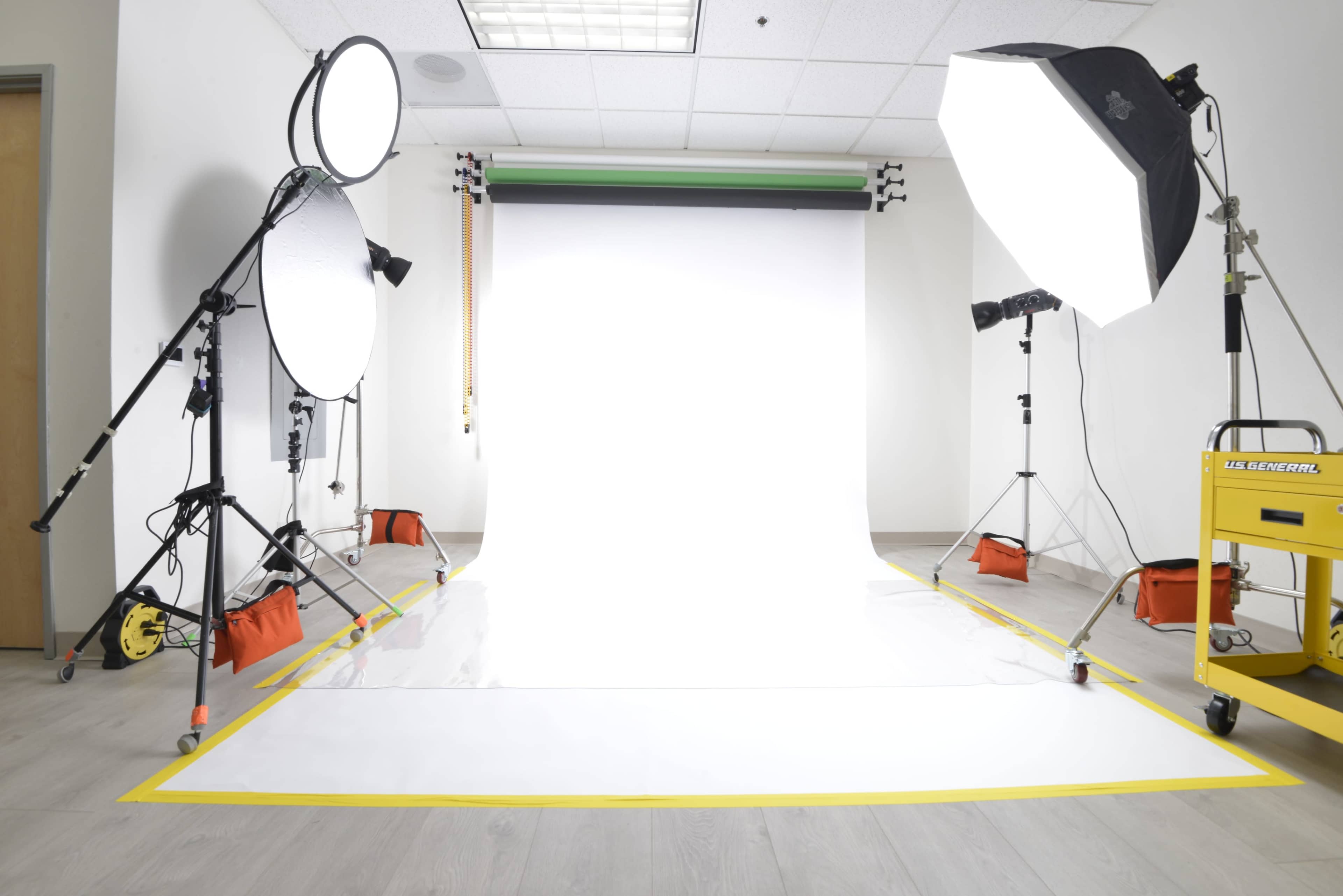 The image shows a photography studio with a white backdrop, multiple lighting equipment, and reflectors set up for a shoot.
