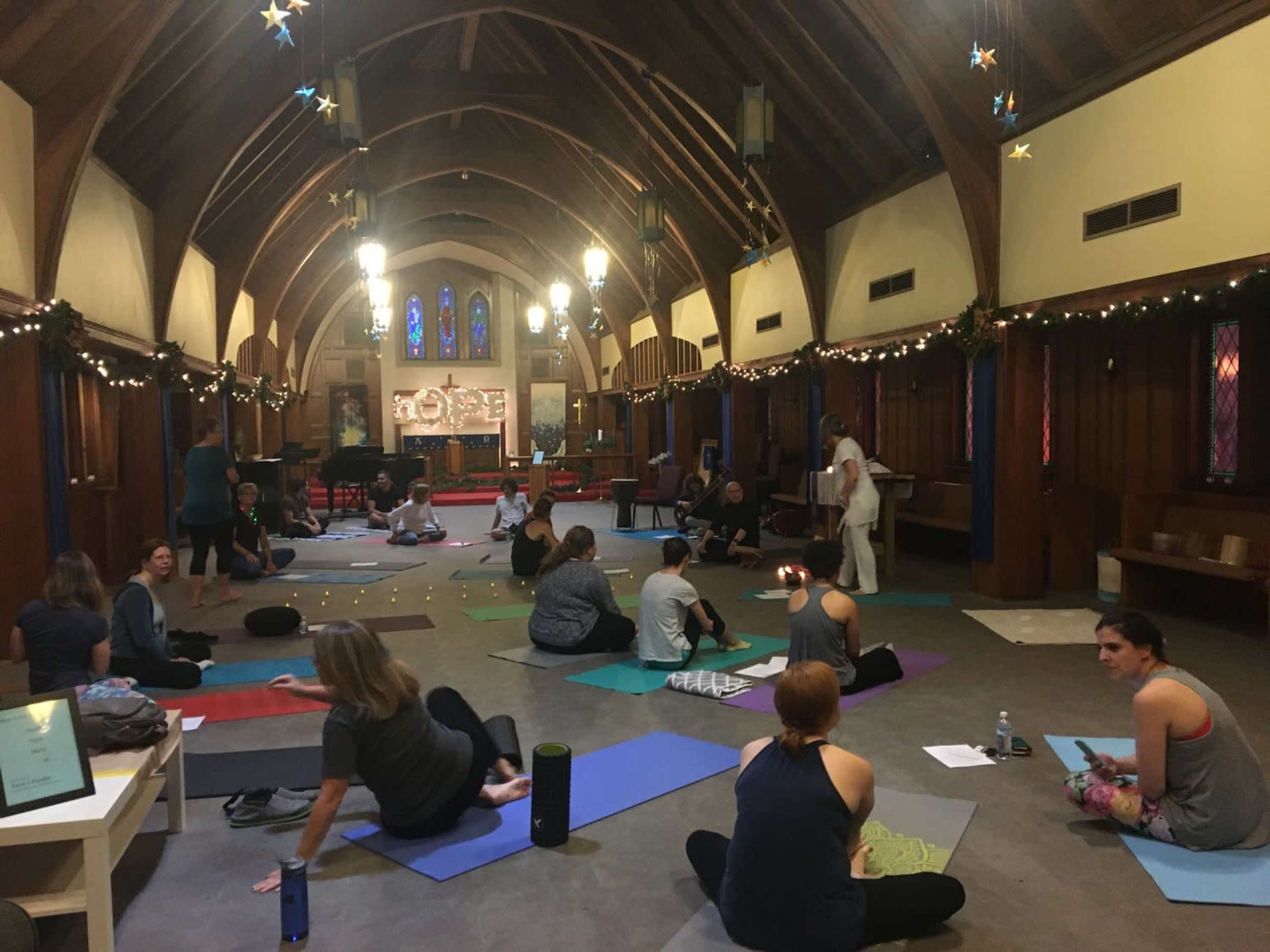 A group of people is engaged in a yoga session inside a spacious, dimly lit room with a high wooden ceiling and decorated with string lights.