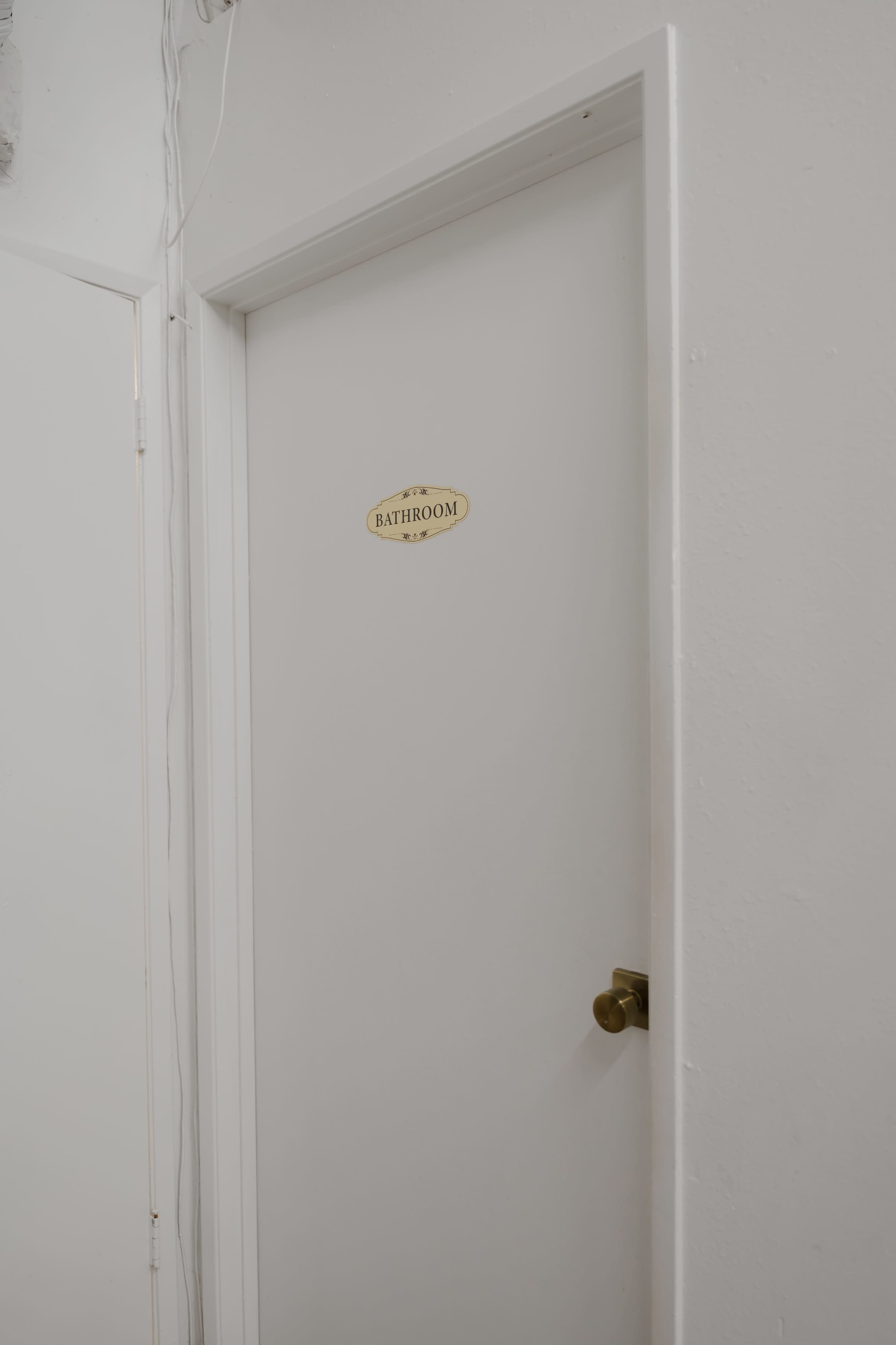 A white door marked with a plaque labeled "BATHROOM" is shown against a plain wall.