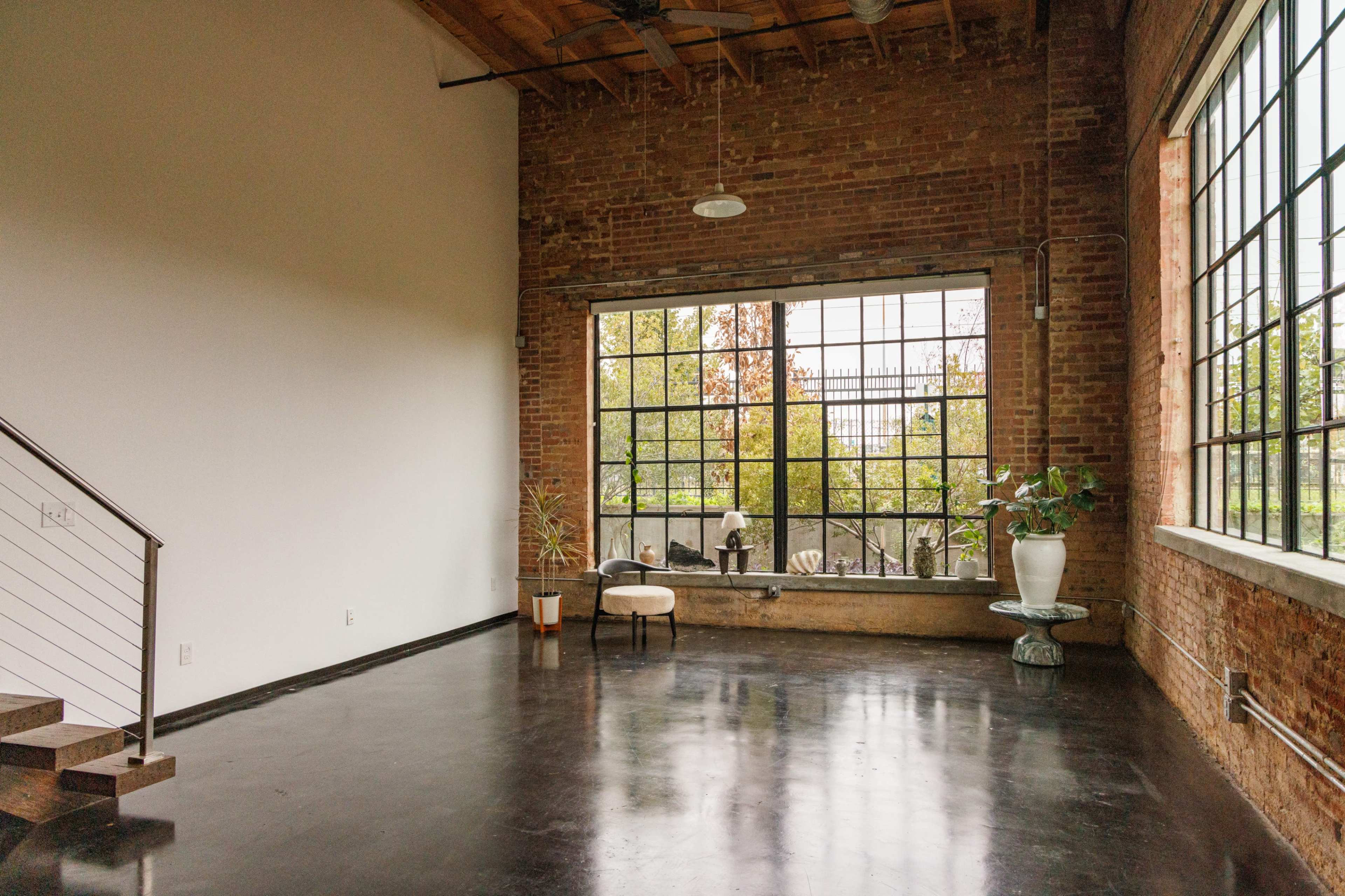 A spacious loft with exposed brick walls, large windows, and polished black flooring contains minimal furniture and indoor plants.