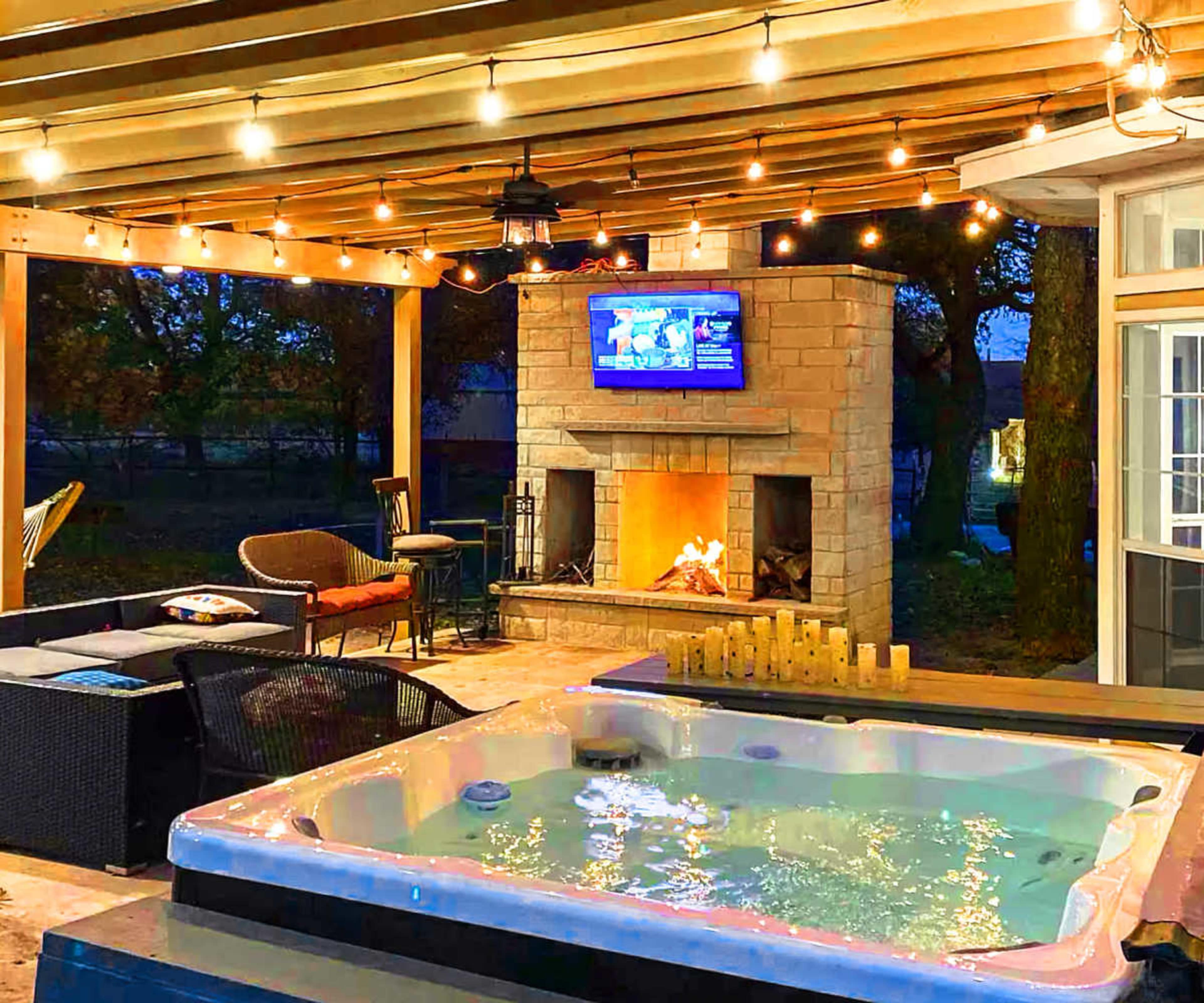 The image shows a covered outdoor patio featuring a hot tub, a stone fireplace with a mounted TV, and ambient string lights.