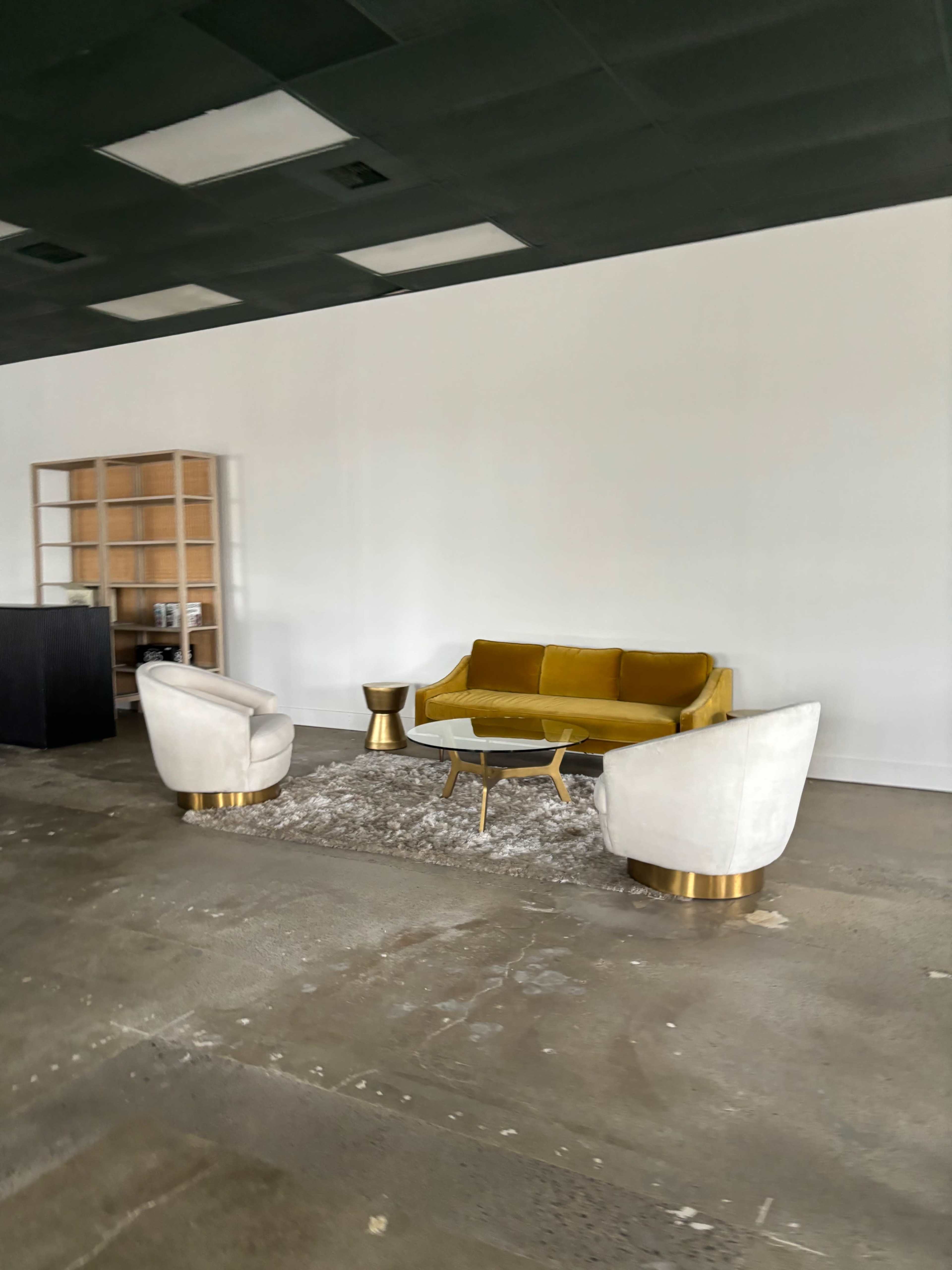 A yellow sofa and two white armchairs are arranged around a small table on a carpet in a sparse, empty room with a concrete floor.
