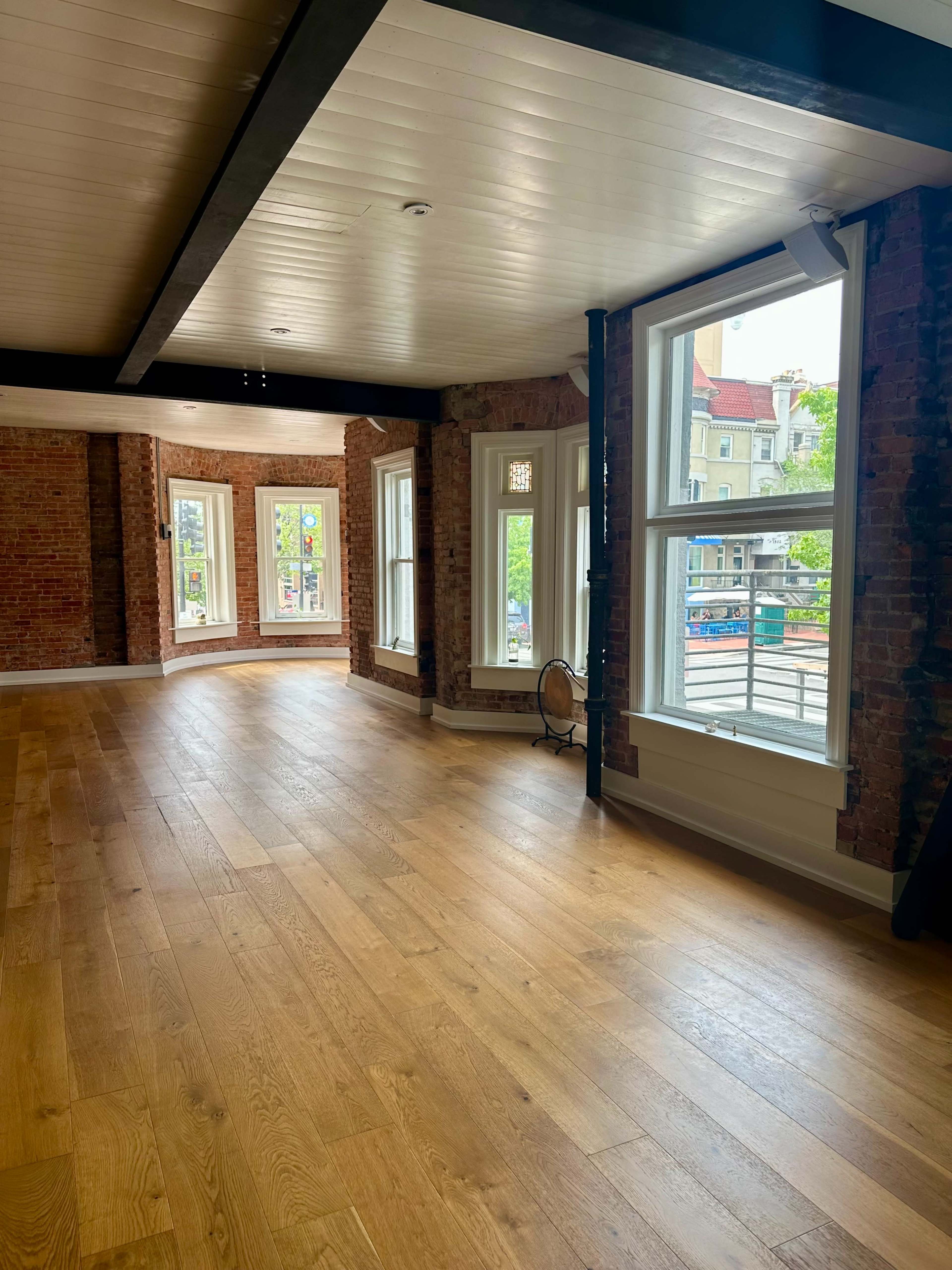 The image shows a spacious interior room with hardwood flooring, exposed brick walls, and large windows allowing natural light to enter.