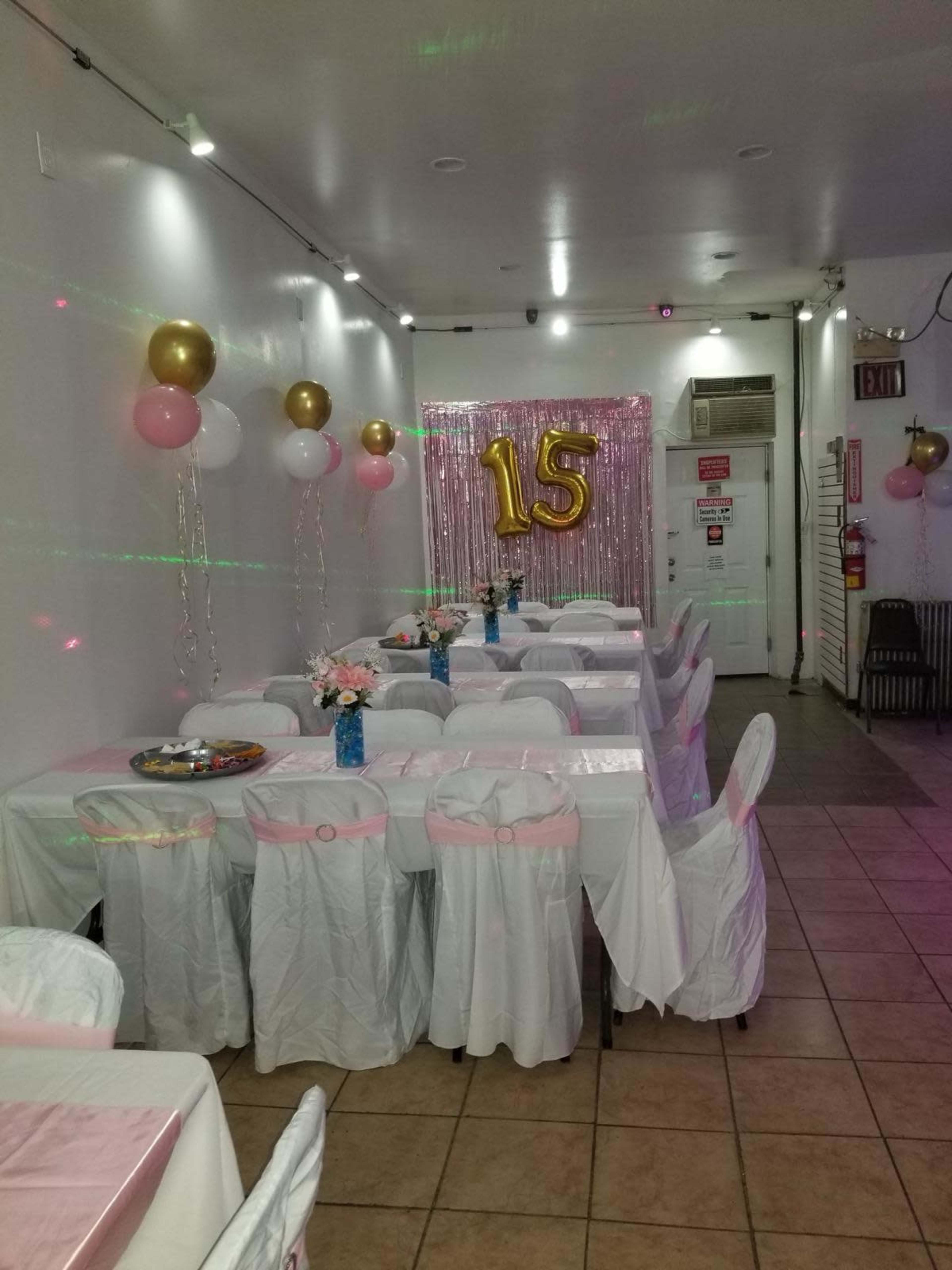 A decorated event space with tables covered in white tablecloths, pink and gold balloons, and a large "15" displayed against a shimmering backdrop.