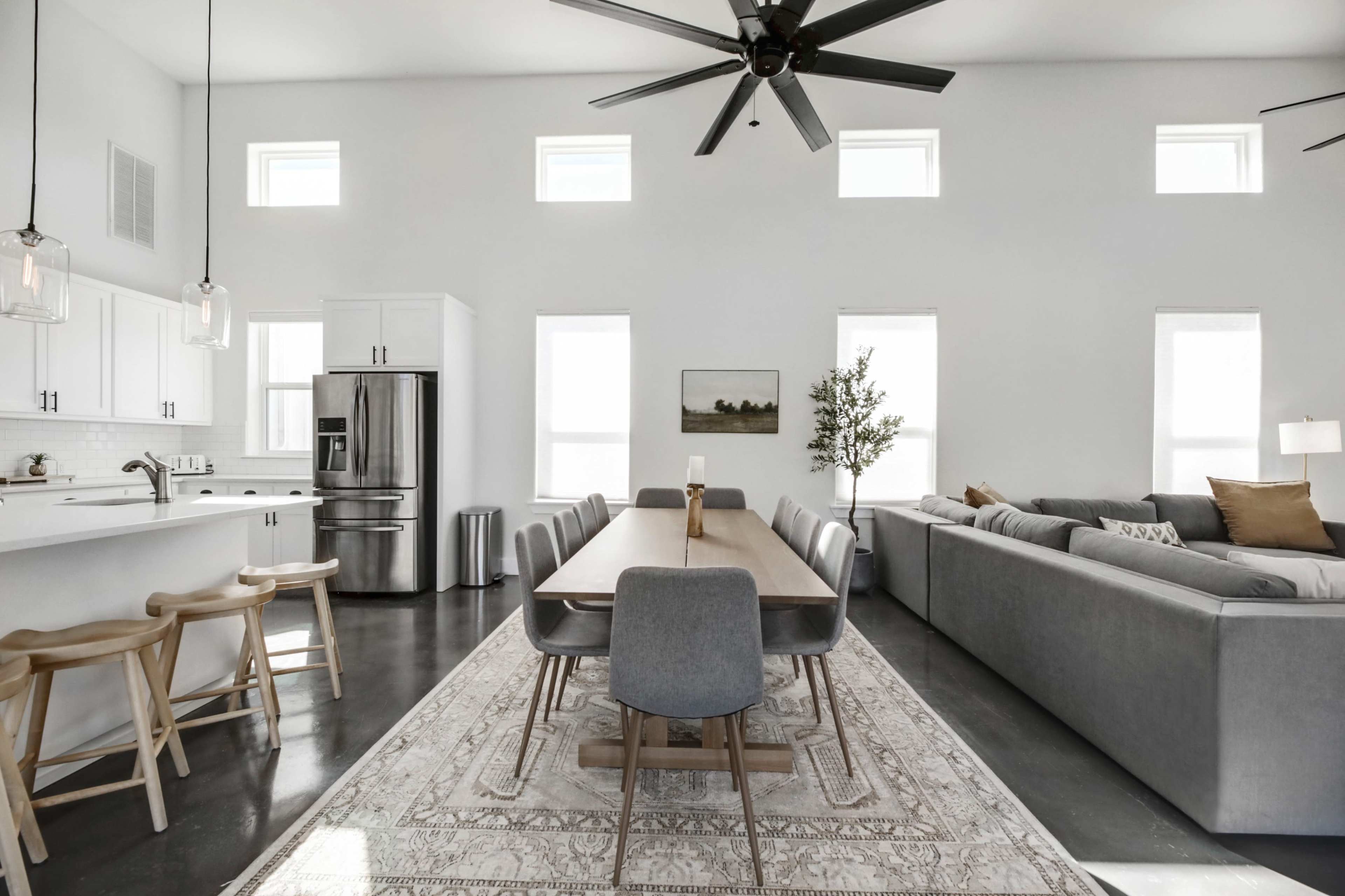 A spacious, modern living area with a dining table, chairs, a kitchen area, and a large sectional sofa, all arranged under a high ceiling with large windows.