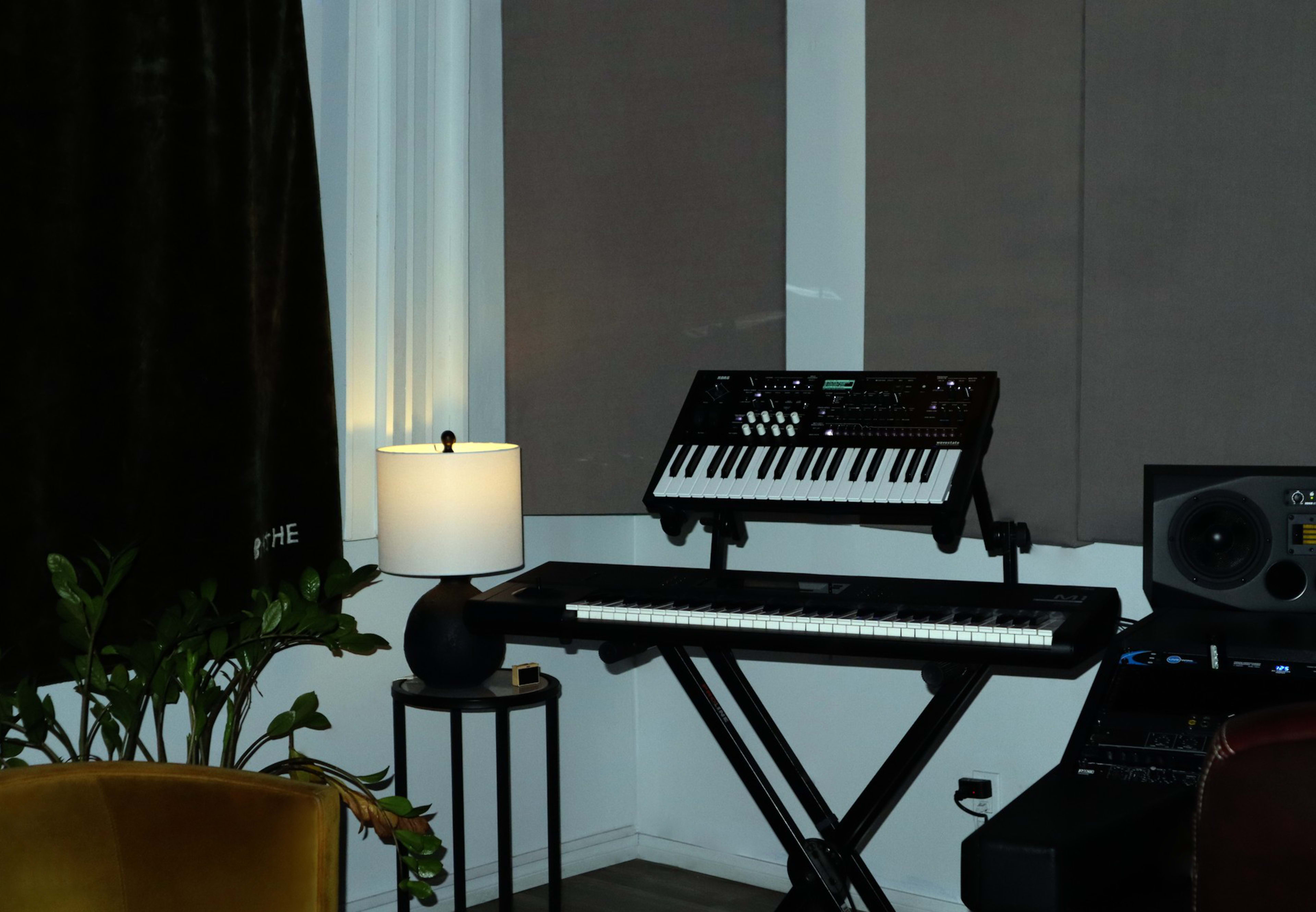 The image shows a modern corner of a room featuring a synthesizer on a stand, a keyboard on a black workstation, a lamp on a side table, and a potted plant.