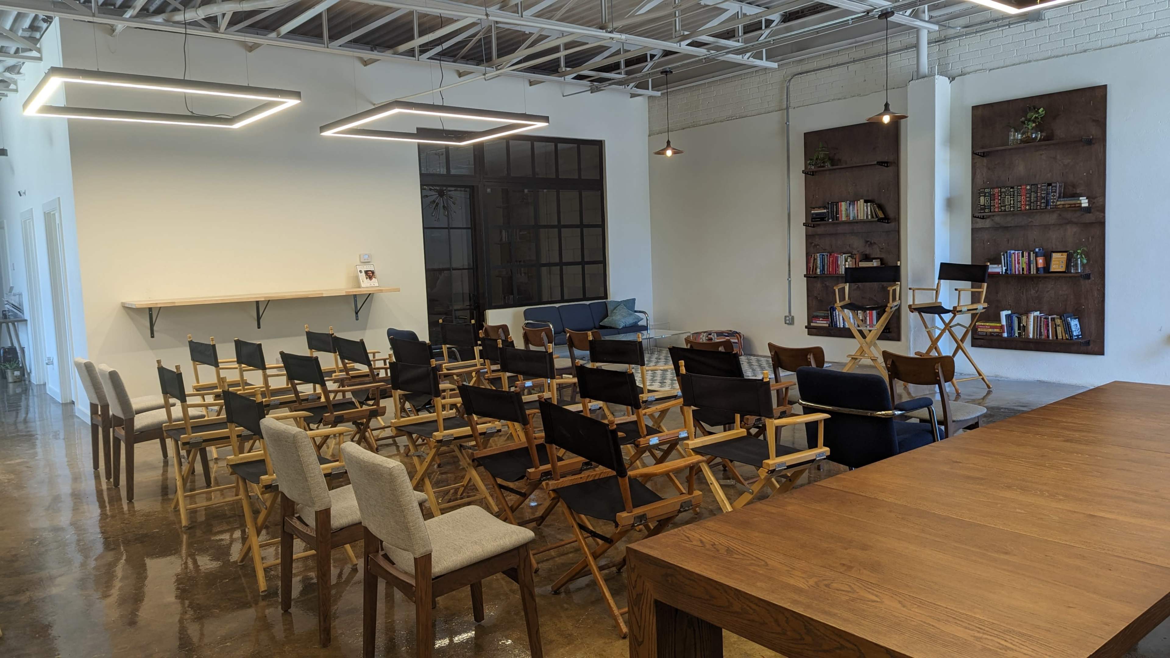 A spacious room with rows of wooden and fabric chairs arranged facing a wall, with bookshelves and a large wooden table along one side.