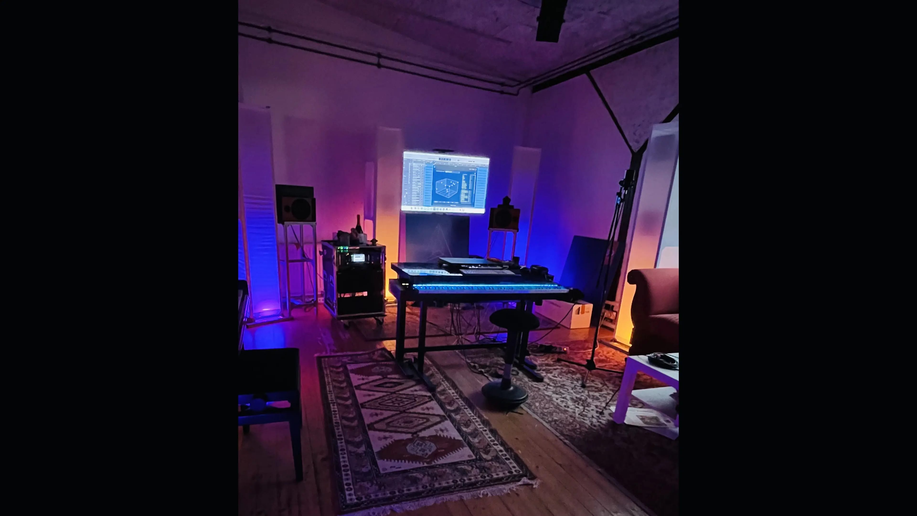 The image shows a music production studio with a central table, electronic equipment, a computer screen displaying music software, and ambient lighting.