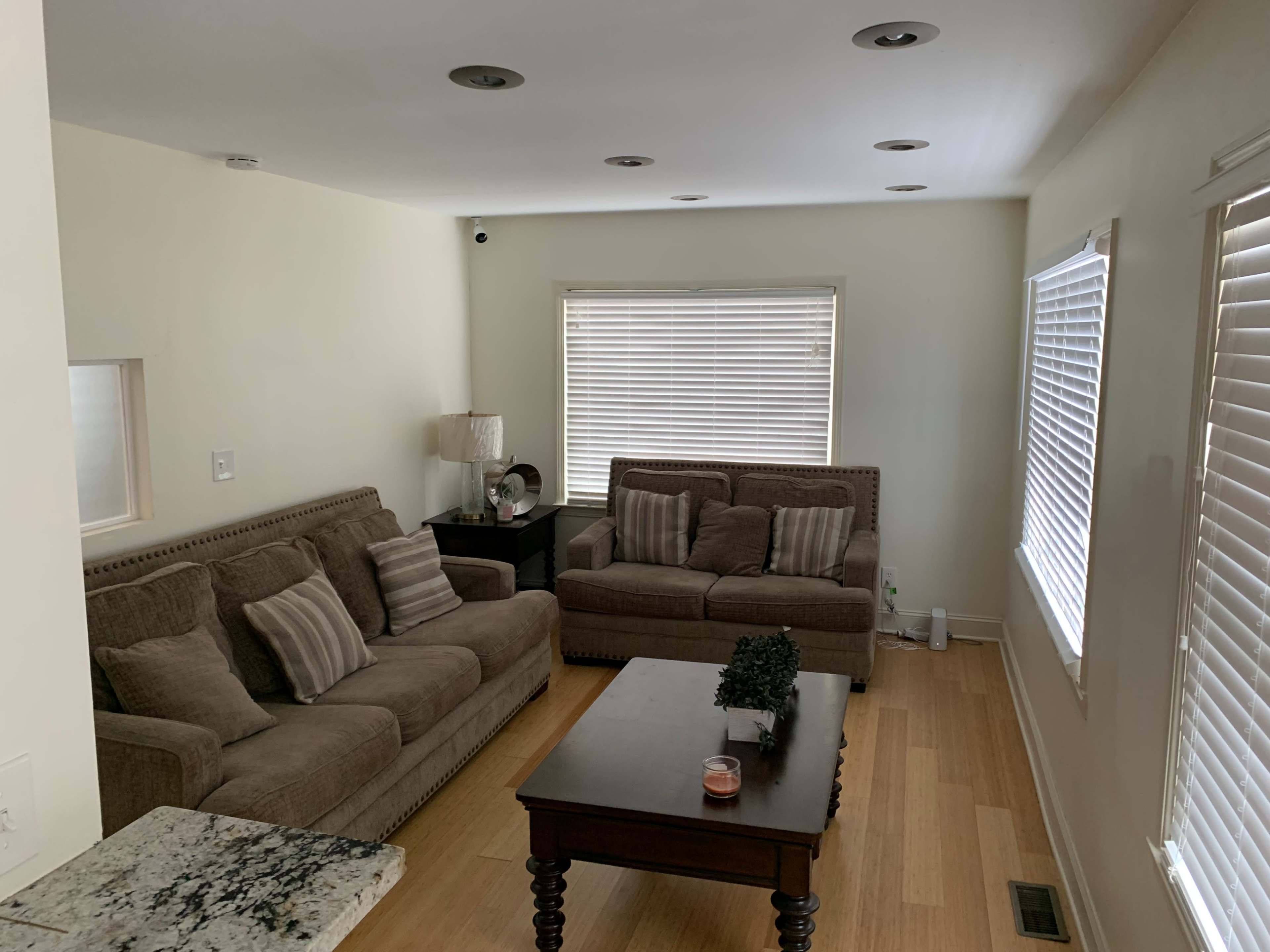 The image shows a living room with two brown sofas, a wooden coffee table, and large windows with white blinds.