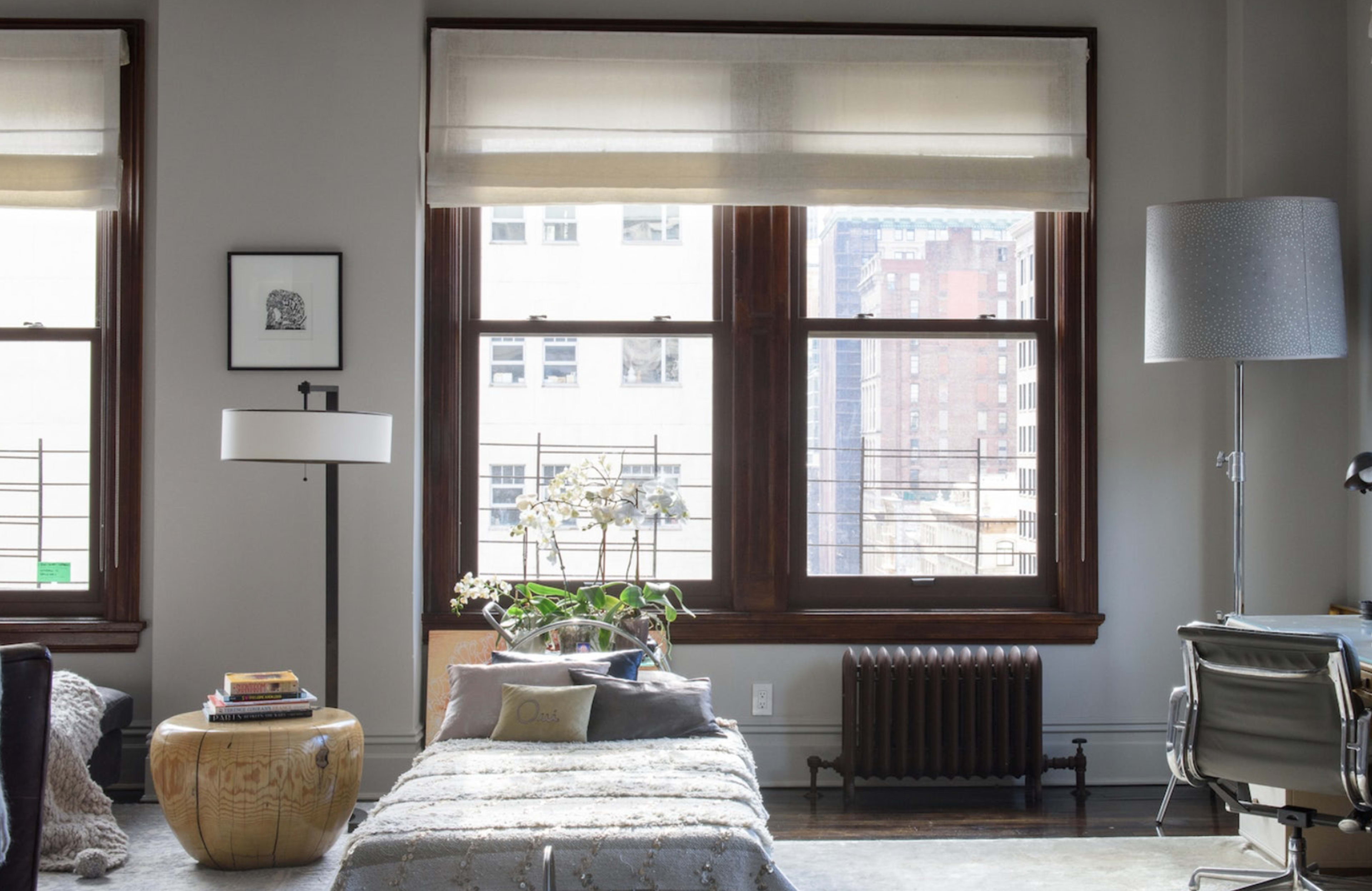 A modern living room features large windows with sheer shades, a wooden coffee table, a radiator, and a desk with a chair, overlooking a cityscape.
