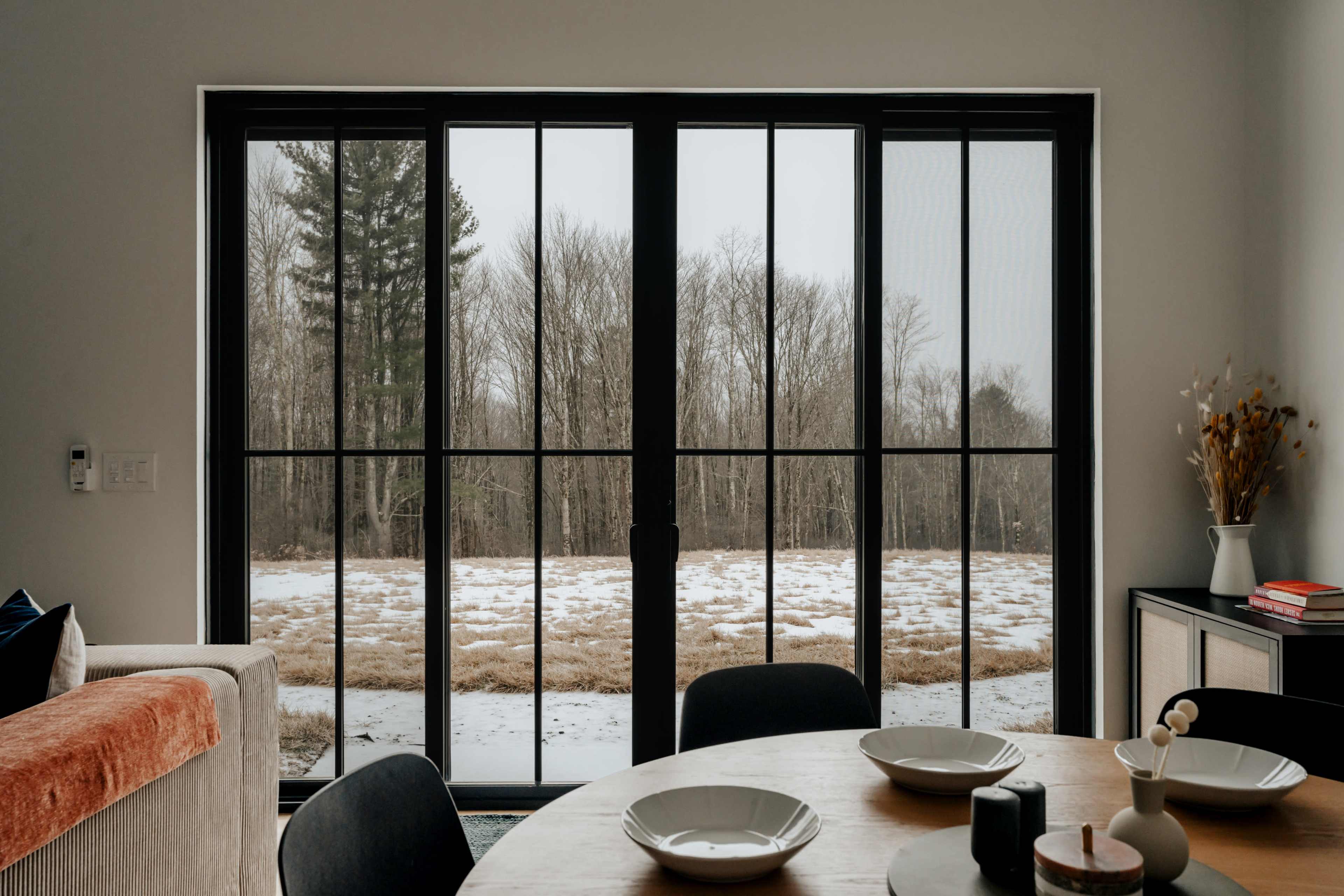 A dining area features a round table set with plates, overlooking a large window that reveals a snowy landscape with bare trees.