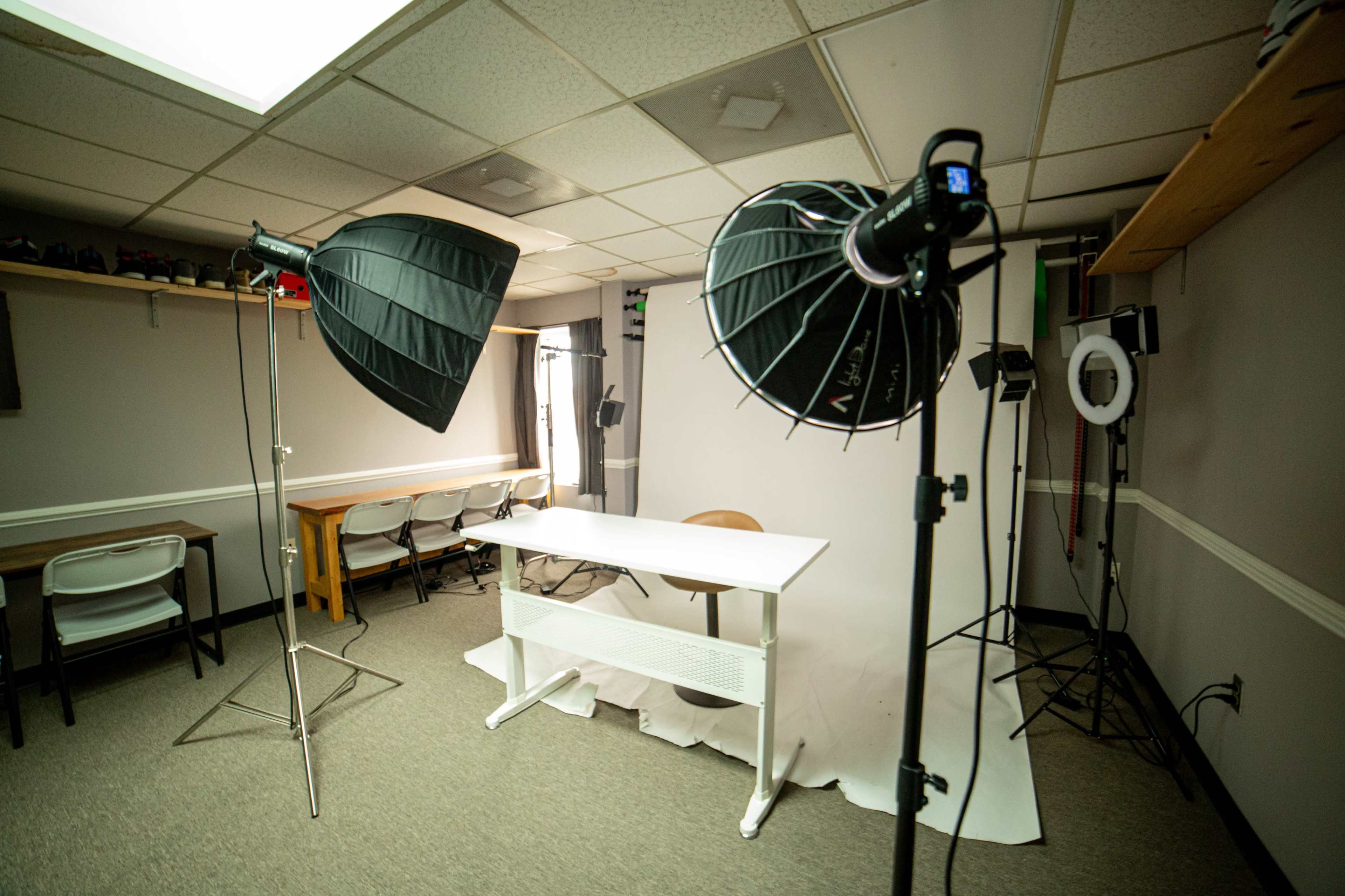 A photography studio setup features a white backdrop, a table with a chair, and multiple lighting equipment on either side.