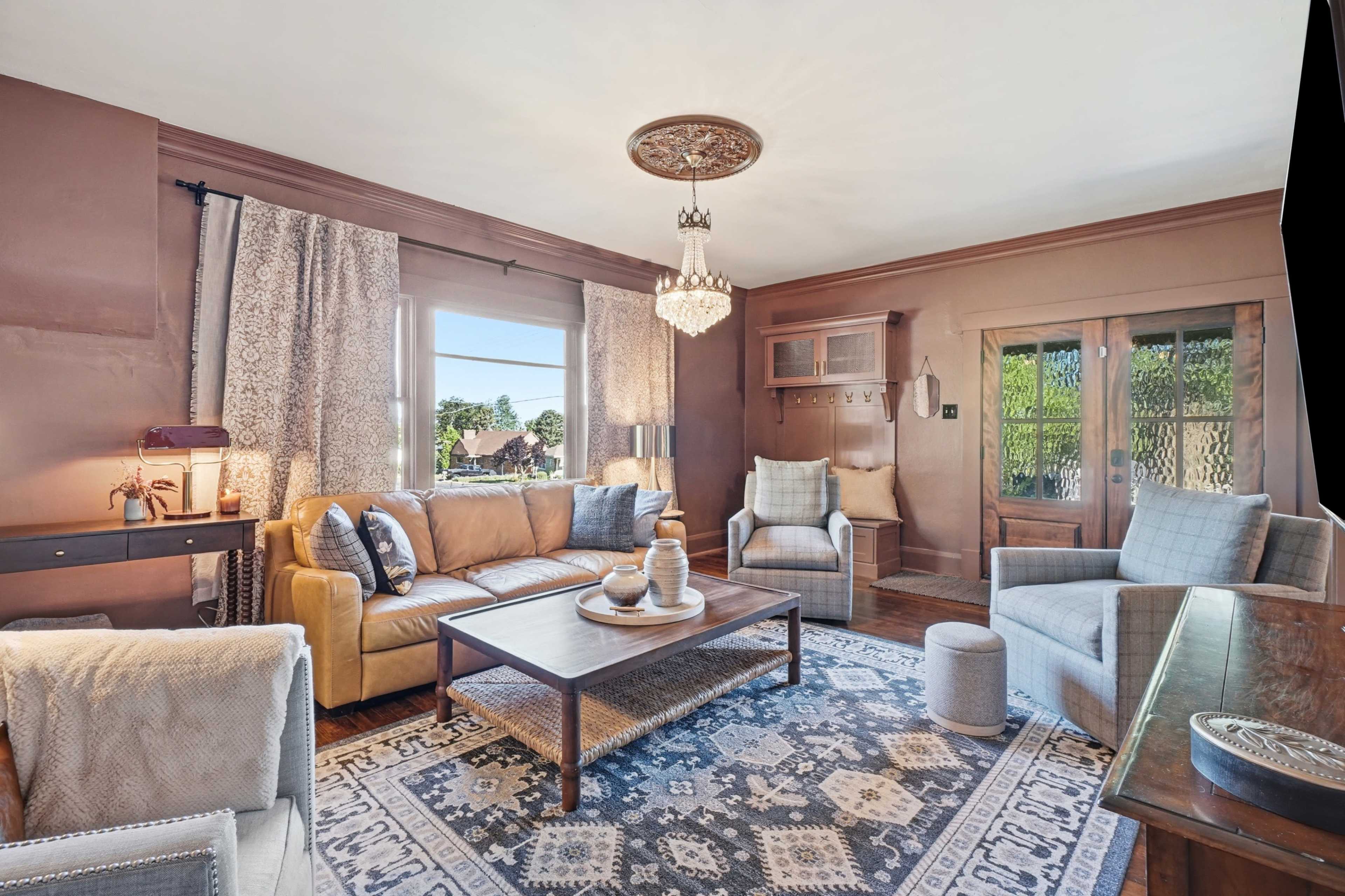 A living room features a brown leather sofa, a wooden coffee table, and patterned curtains, with light streaming in from a large window.