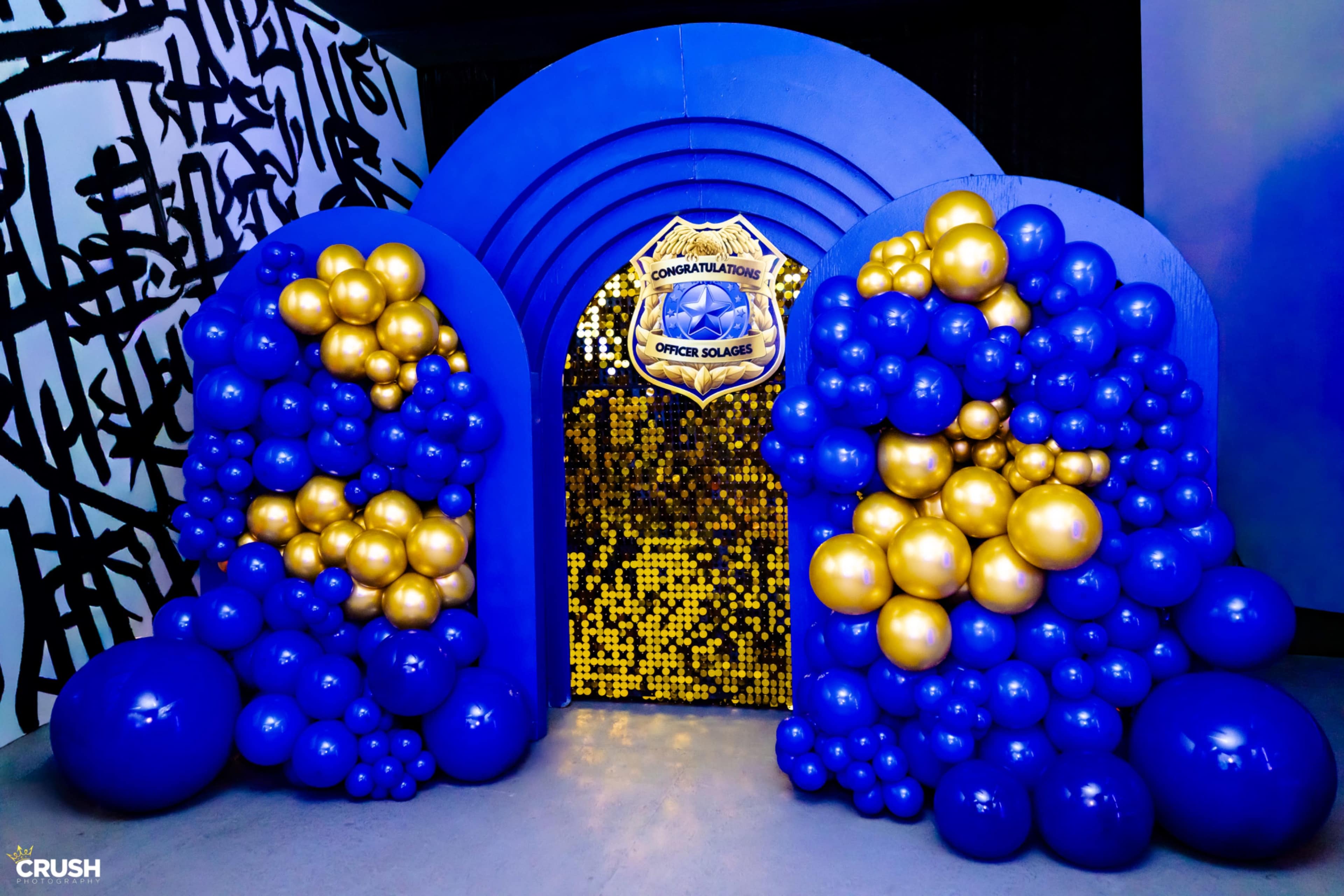 A festive archway made of blue and gold balloons leads to a sequined backdrop, adorned with a badge that reads "Congratulations Officer Squales."