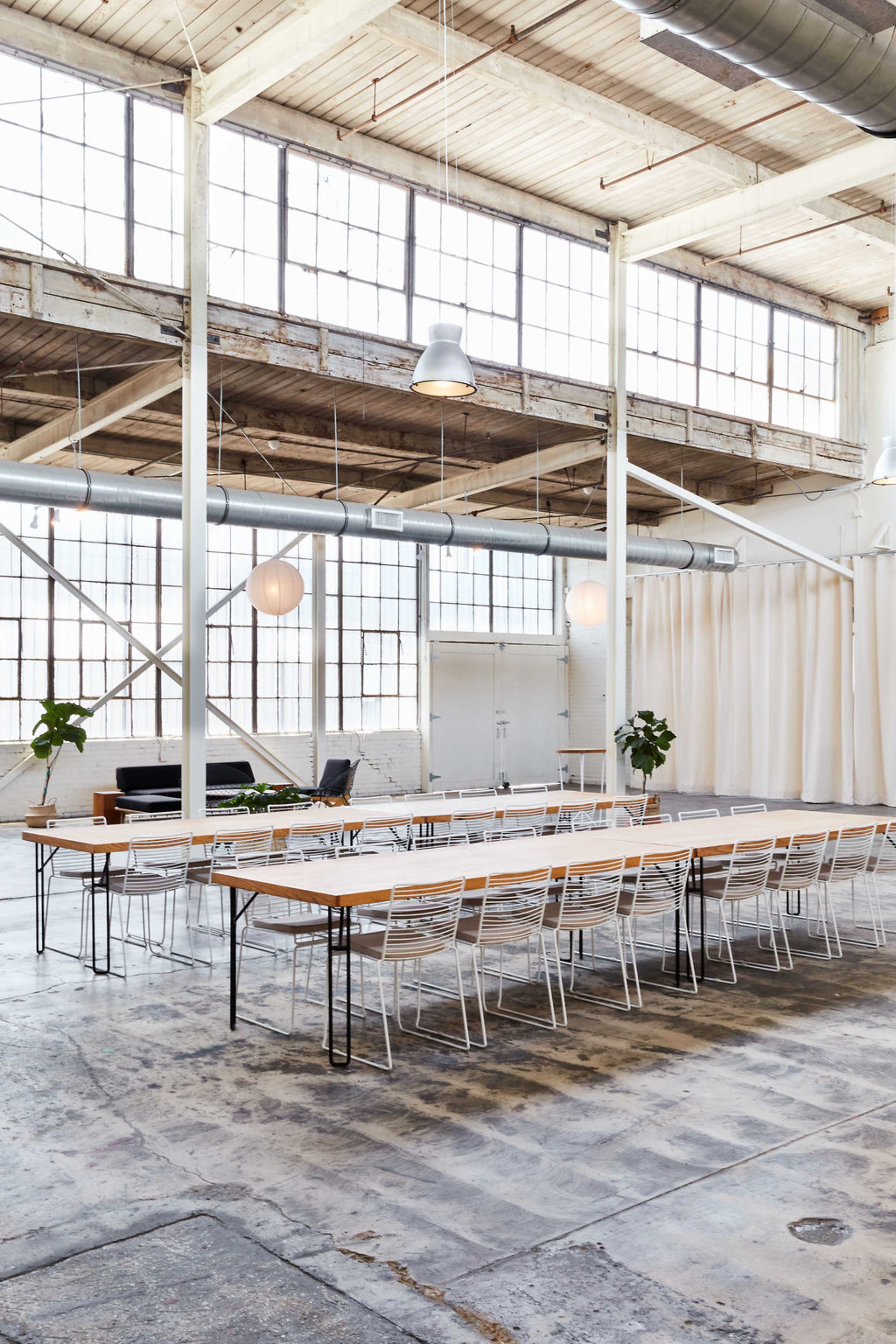 The image shows a spacious, industrial-style room with large windows, a long wooden dining table surrounded by white chairs, and green plants in the corners.
