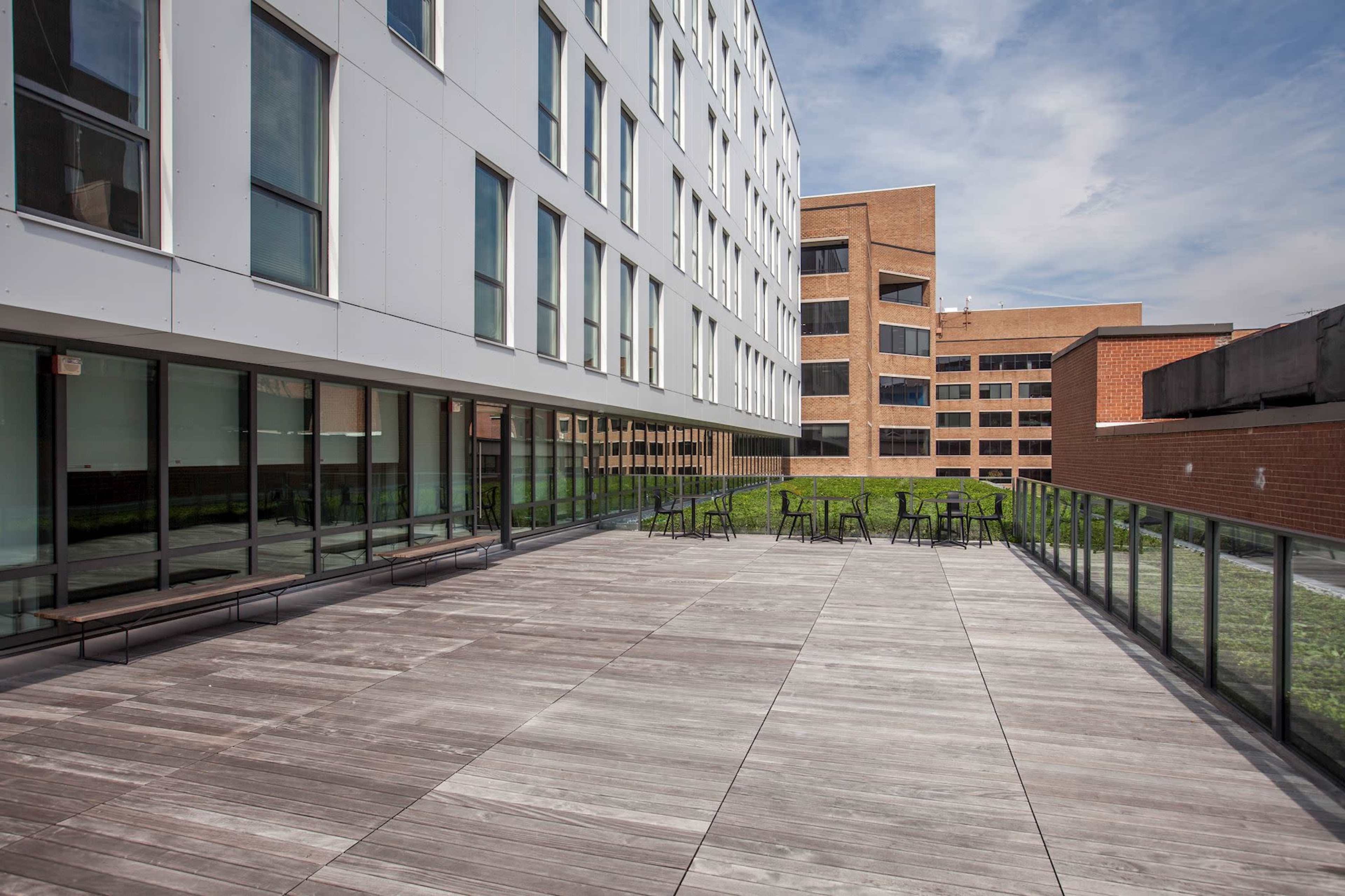 The image shows a spacious outdoor terrace with wooden flooring, furnished with metal tables and chairs, surrounded by modern buildings.