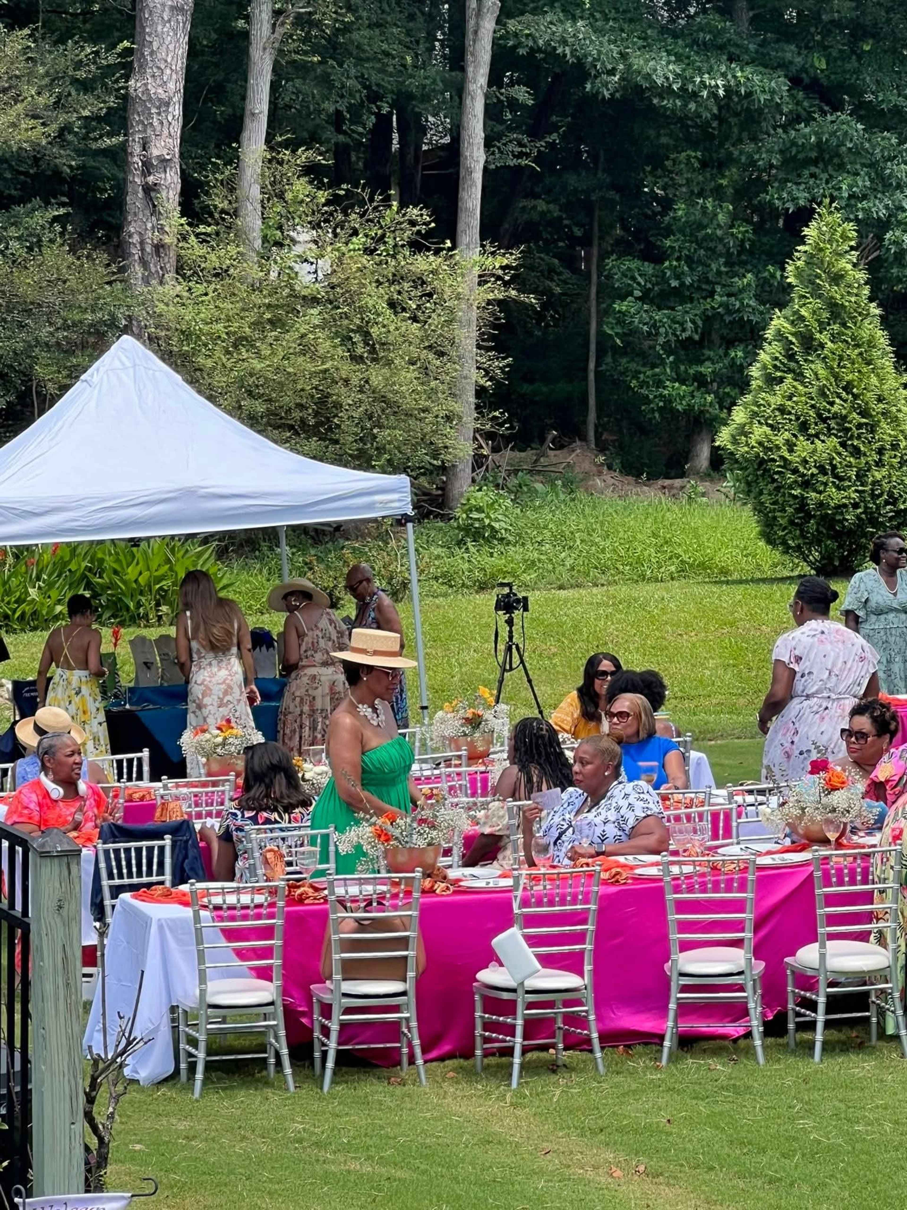 A large outdoor gathering features numerous guests seated at tables draped in pink cloth, with a white tent in the background and a lush green landscape.