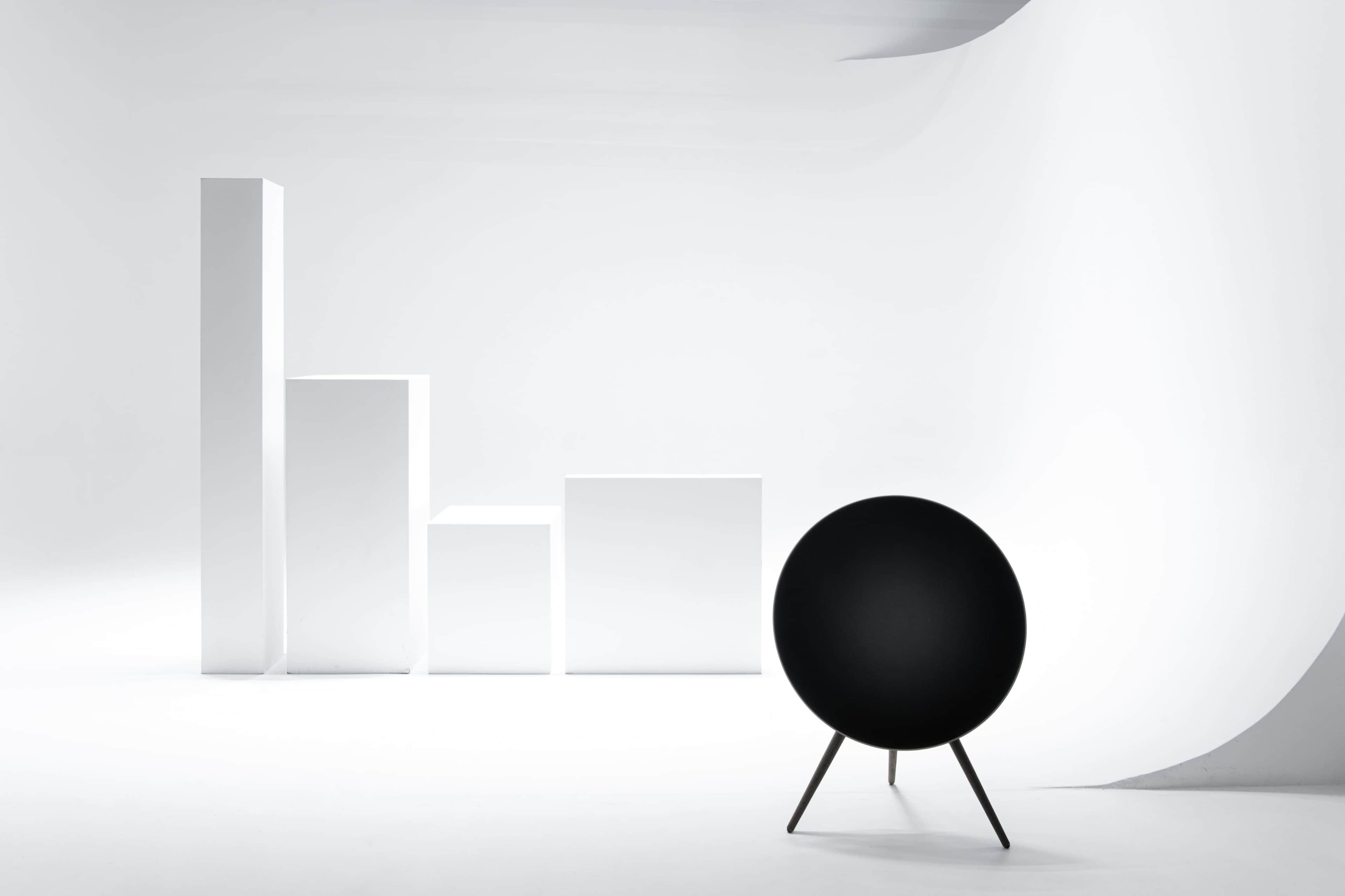 The image features a black circular speaker on a tripod next to three white rectangular blocks arranged in varying heights against a minimalist white background.