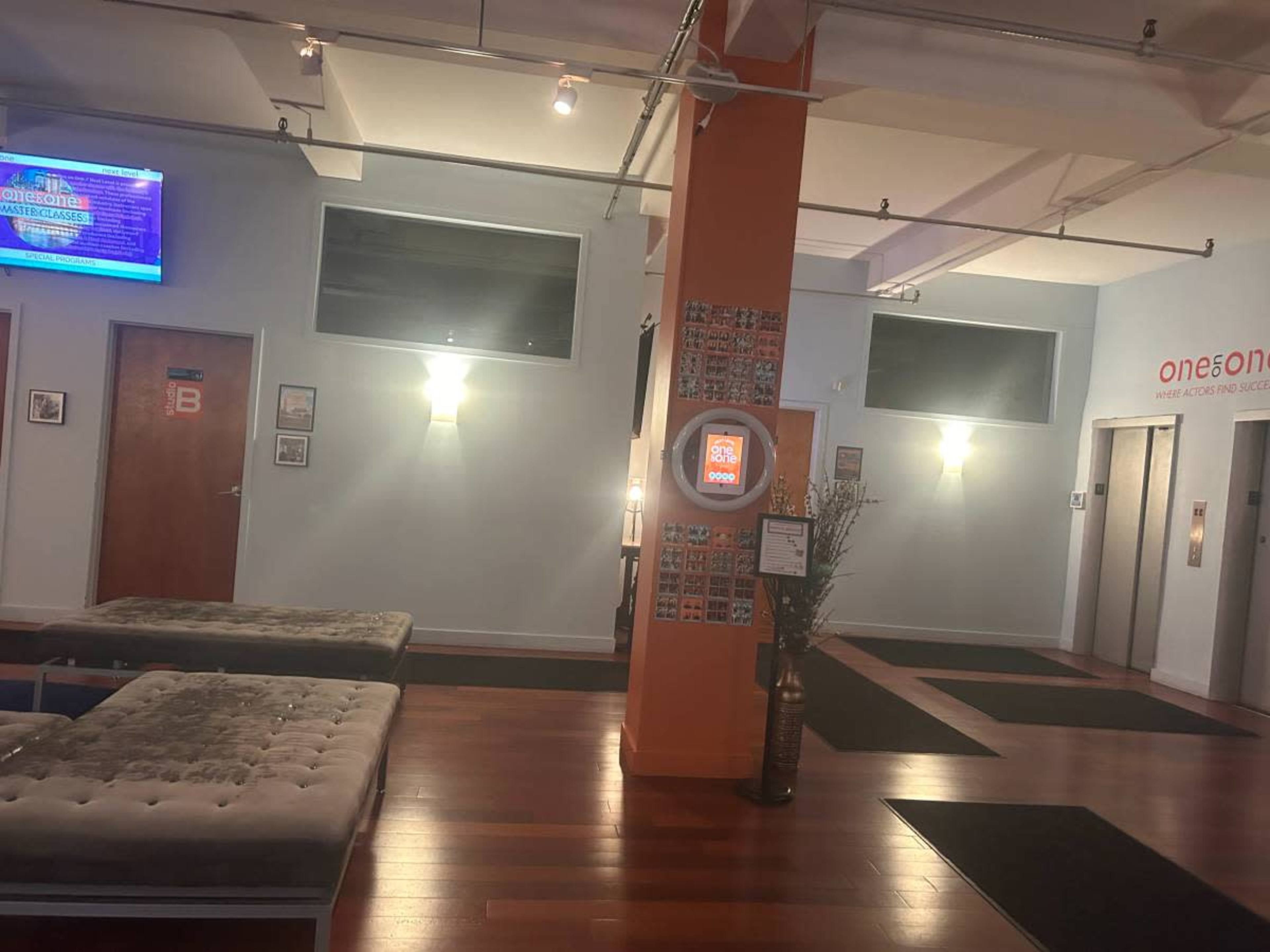 The image shows a modern lobby area with wooden flooring, a seating arrangement, an elevator, and walls adorned with framed photos.