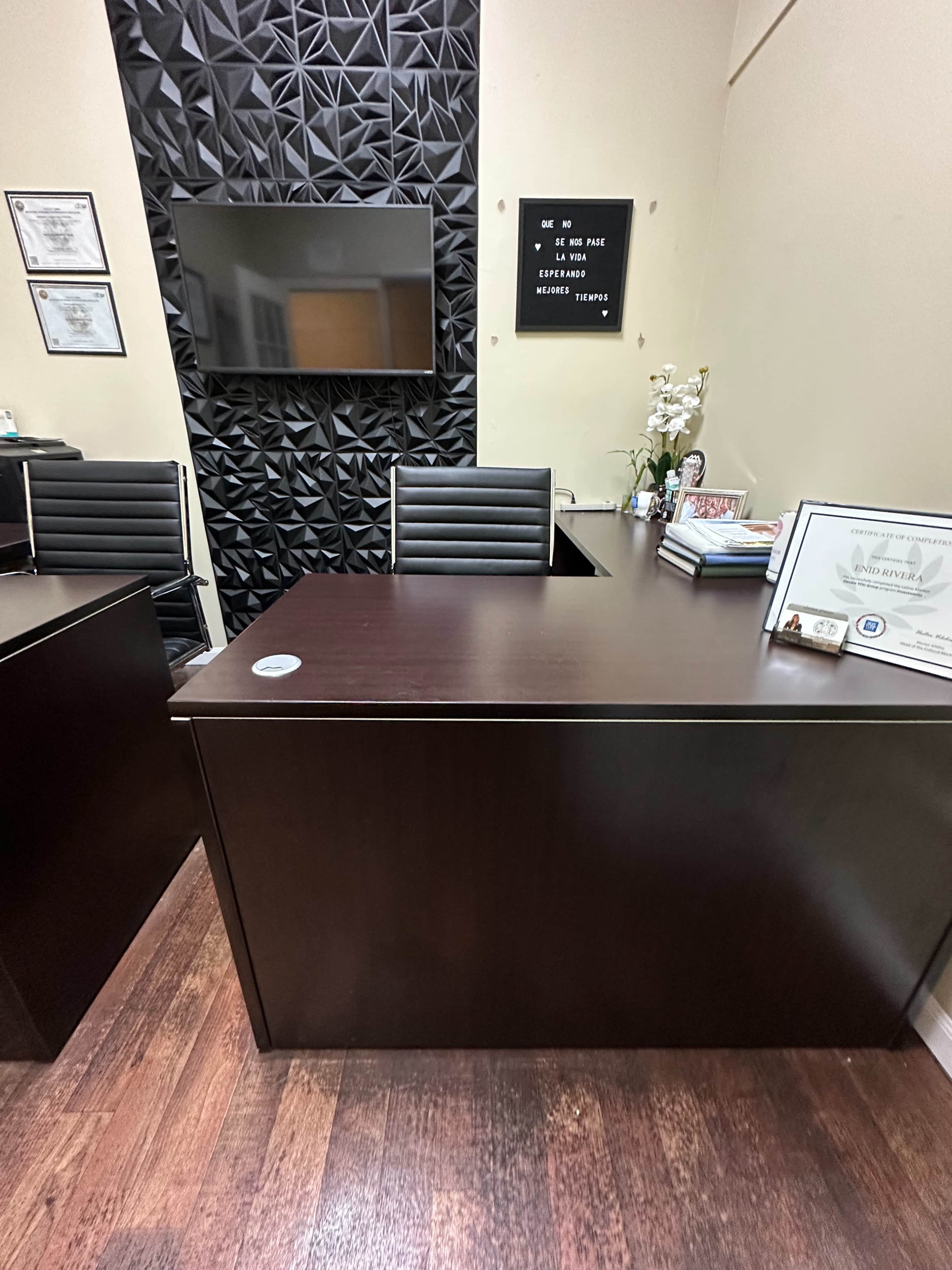 The image shows a modern office space with two dark wood desks, a wall featuring a textured black panel, and a television mounted above, alongside framed certificates and a potted plant on one desk.